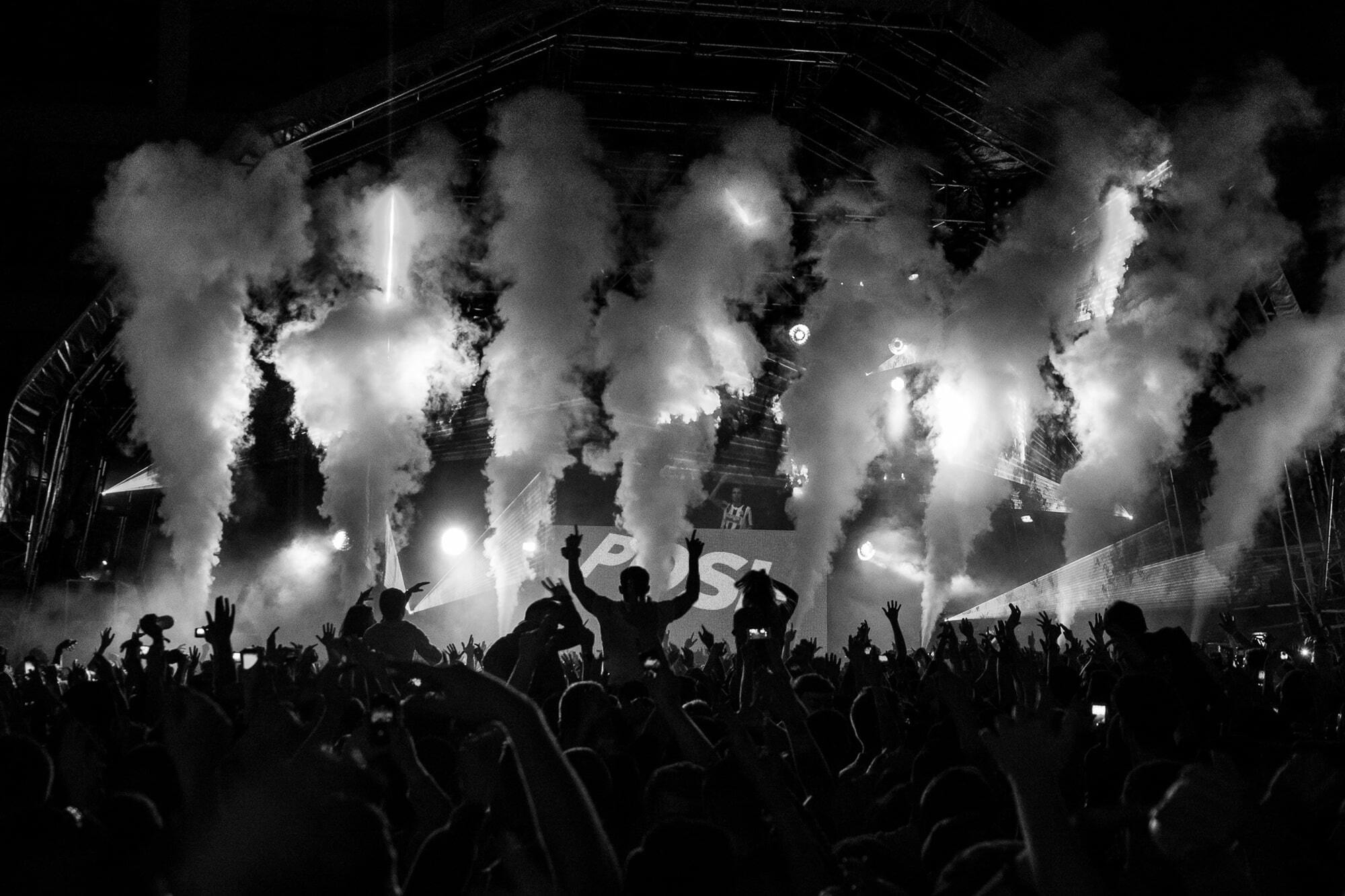 Black-and-white concert crowd raising hands as CO2 jets erupt over a Posh stage