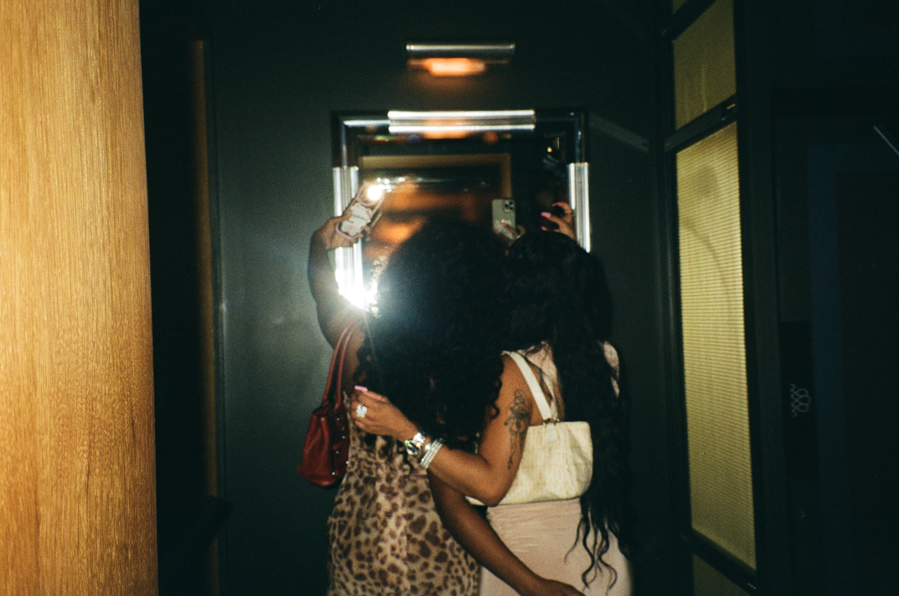 Two women take a flash mirror selfie in a dim hallway with patterned handbags