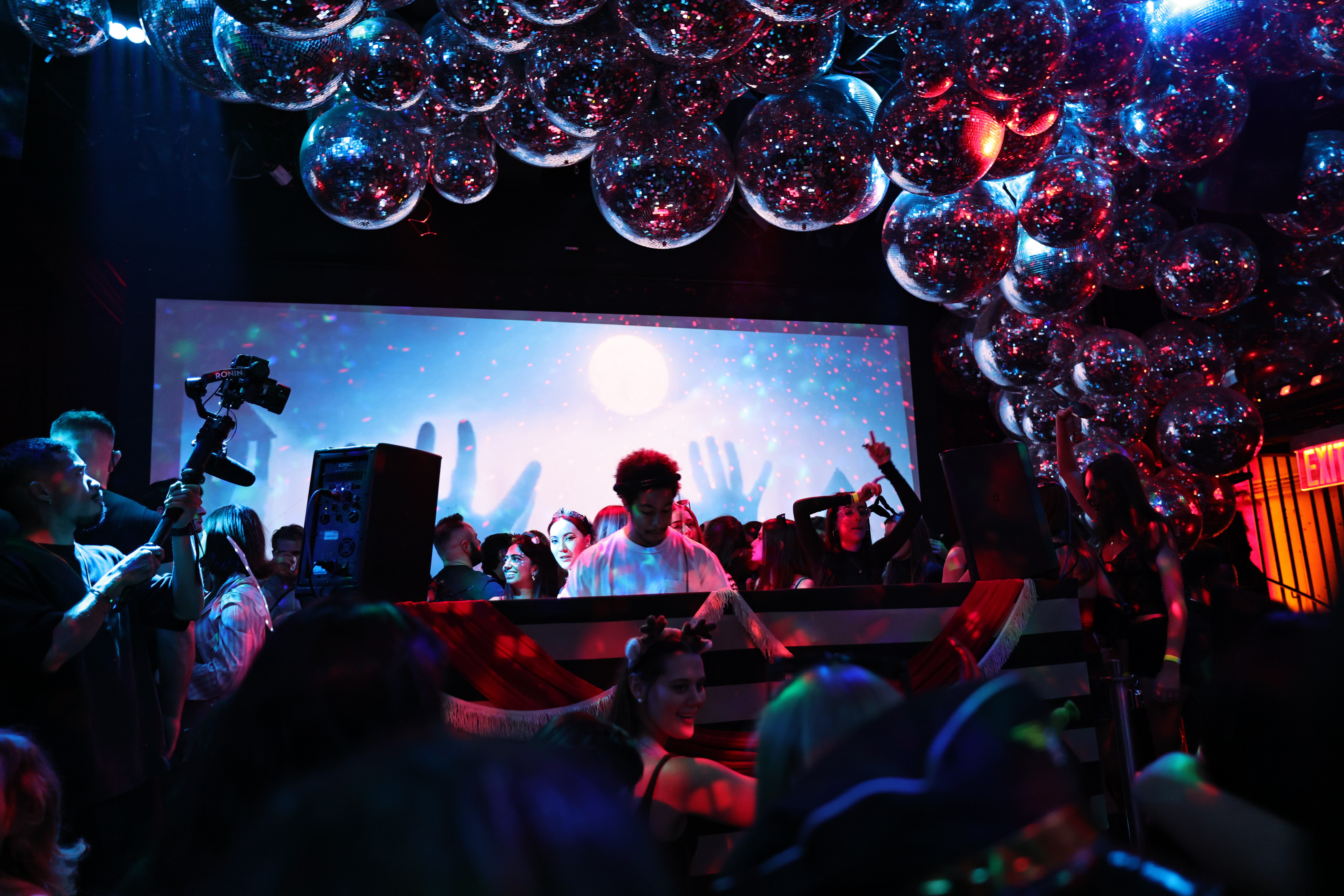 DJ performs under a ceiling of disco balls before a glowing LED screen