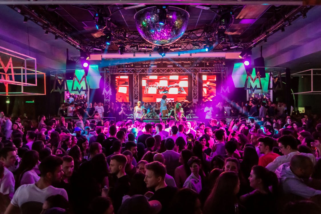 Dense crowd dances under a disco ball before neon 'M' club signage
