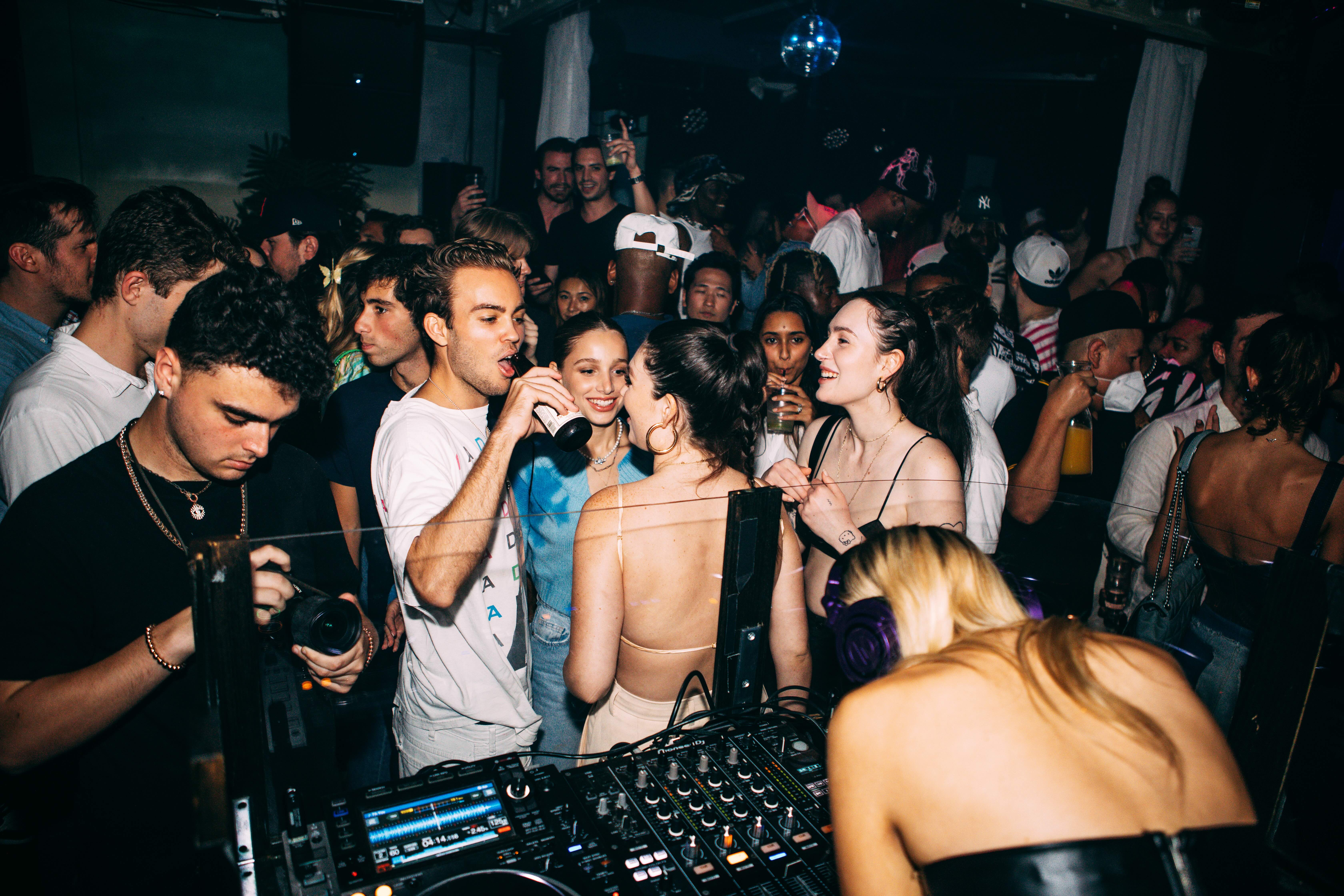 Young partygoers drink and mingle behind a DJ mixer on the dancefloor