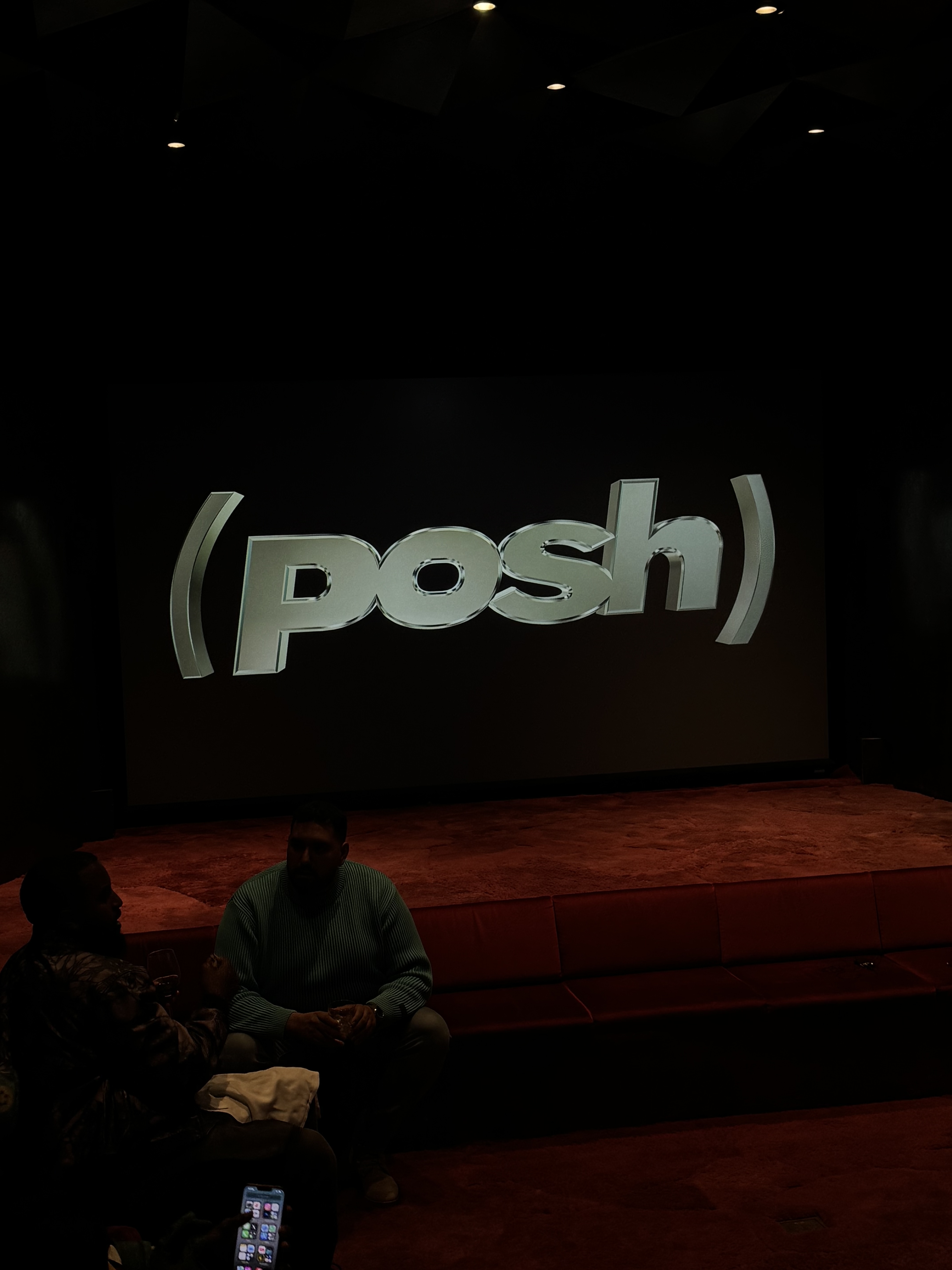 Two men sit on red couches before a large illuminated 'posh' logo screen