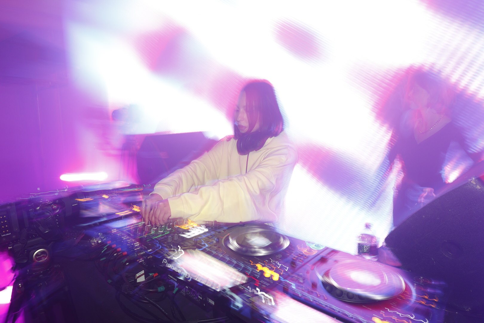 DJ in a white hoodie mixes on Pioneer decks amid blurred purple lights
