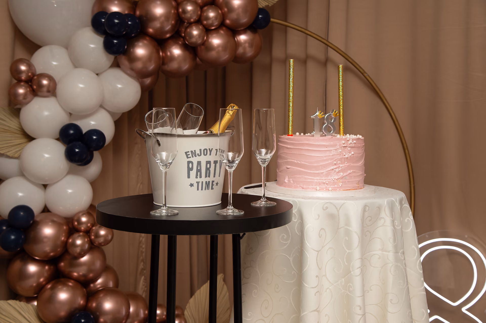 Roze rođendanska torta sa svećicama 18 pored bele kante sa šampanjcem i čašama na crnom stolu, okruženo balonima.