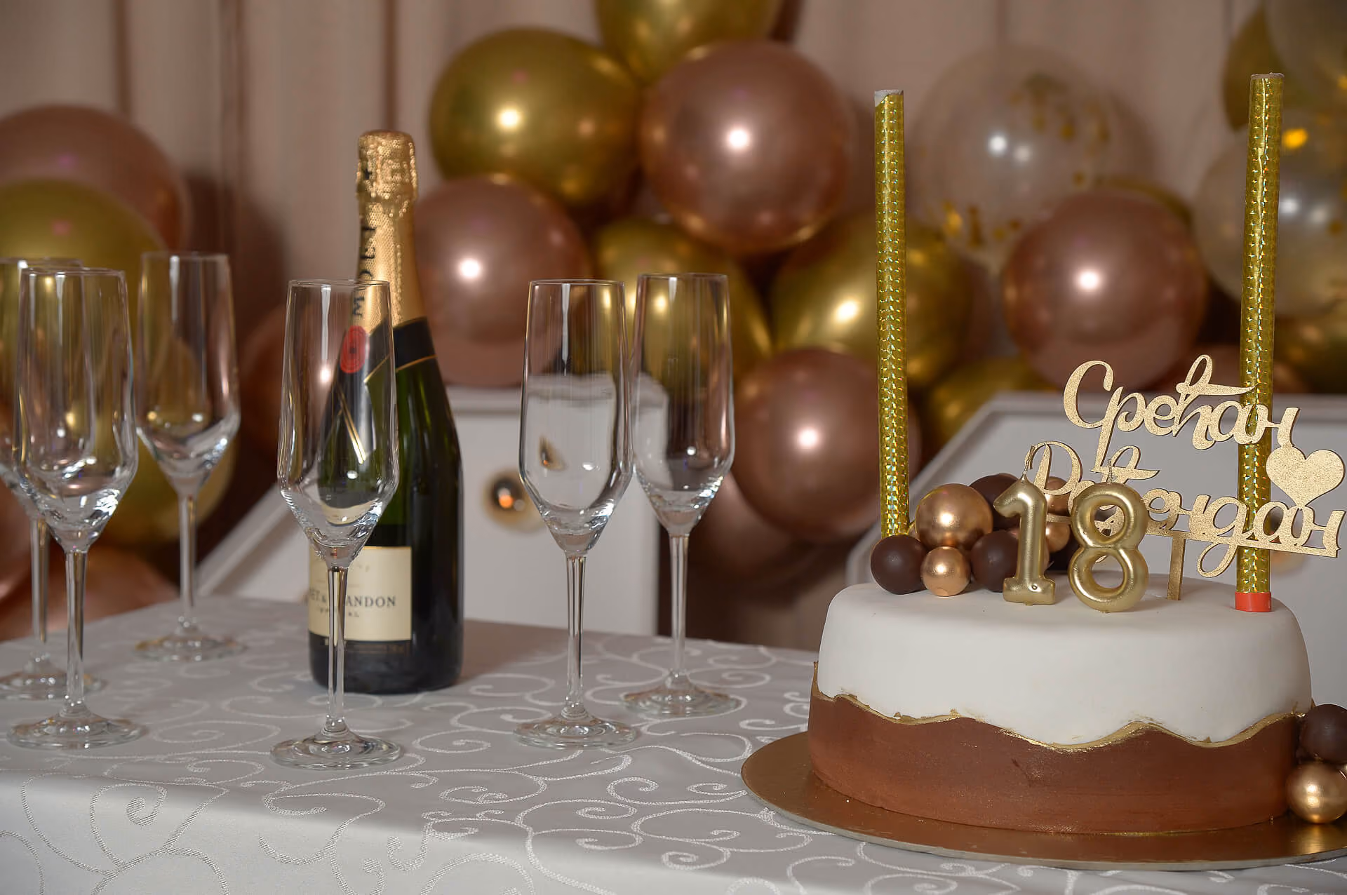Rođendanska torta sa zlatnim brojem 18 i natpisom Srećan Rođendan, pored boca šampanjca i šest praznih čaša na stolu sa belim stolnjakom, i baloni u pozadini.