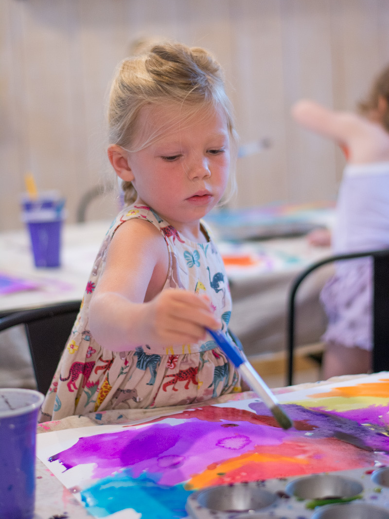 A girl holds a paintbrush, adding purple to an abstract kid's painting.