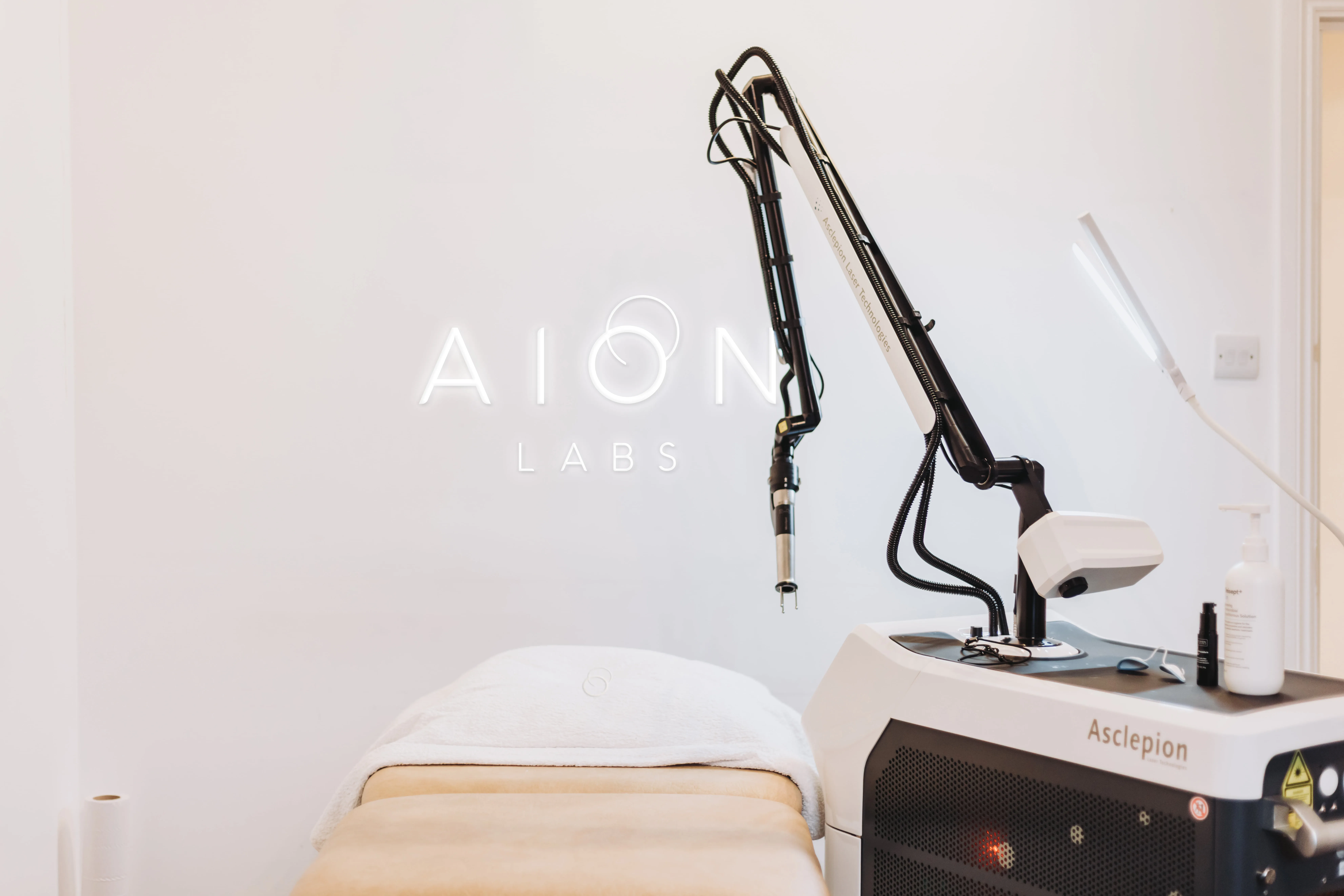 Medical or skincare treatment room with a beige treatment bed and a laser technology machine beside it, under AION LABS neon sign on a white wall.