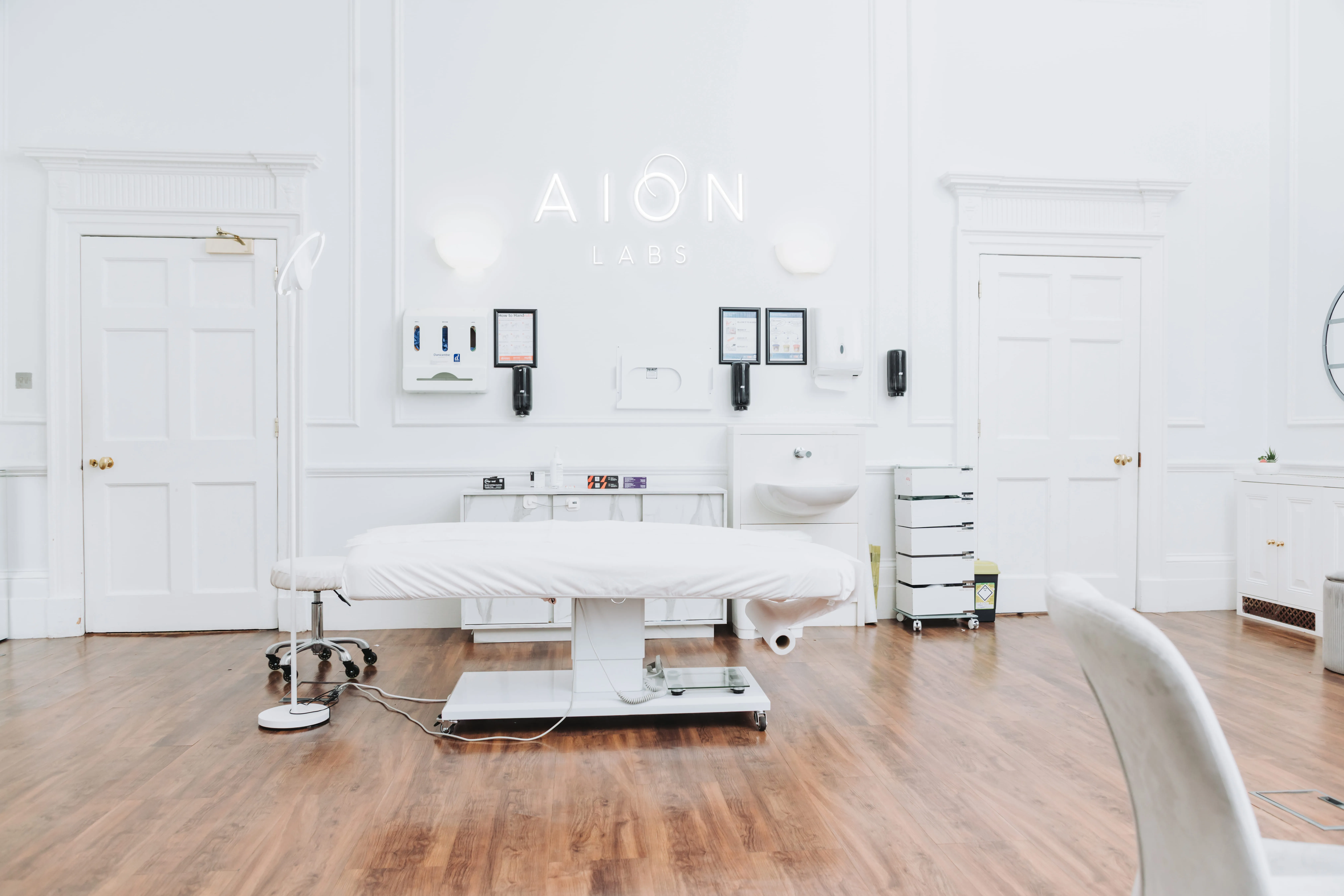 Bright modern medical examination room with a white treatment table, wooden floor, hand sanitizer dispensers, and 'AION LABS' sign on the back wall.