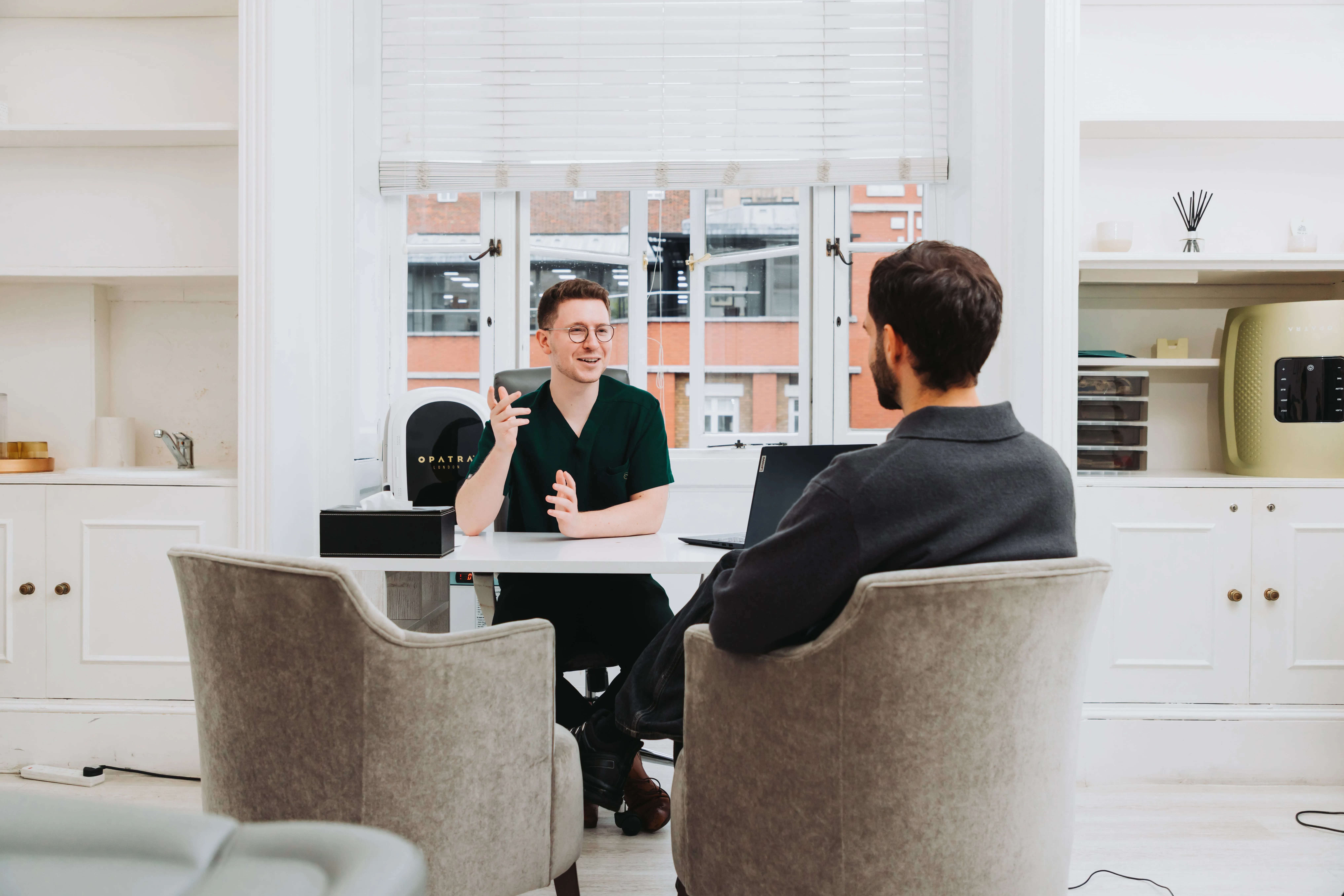 Modern indoor swimming pool with green underwater lights, lounge chairs, potted plants, and large windows.