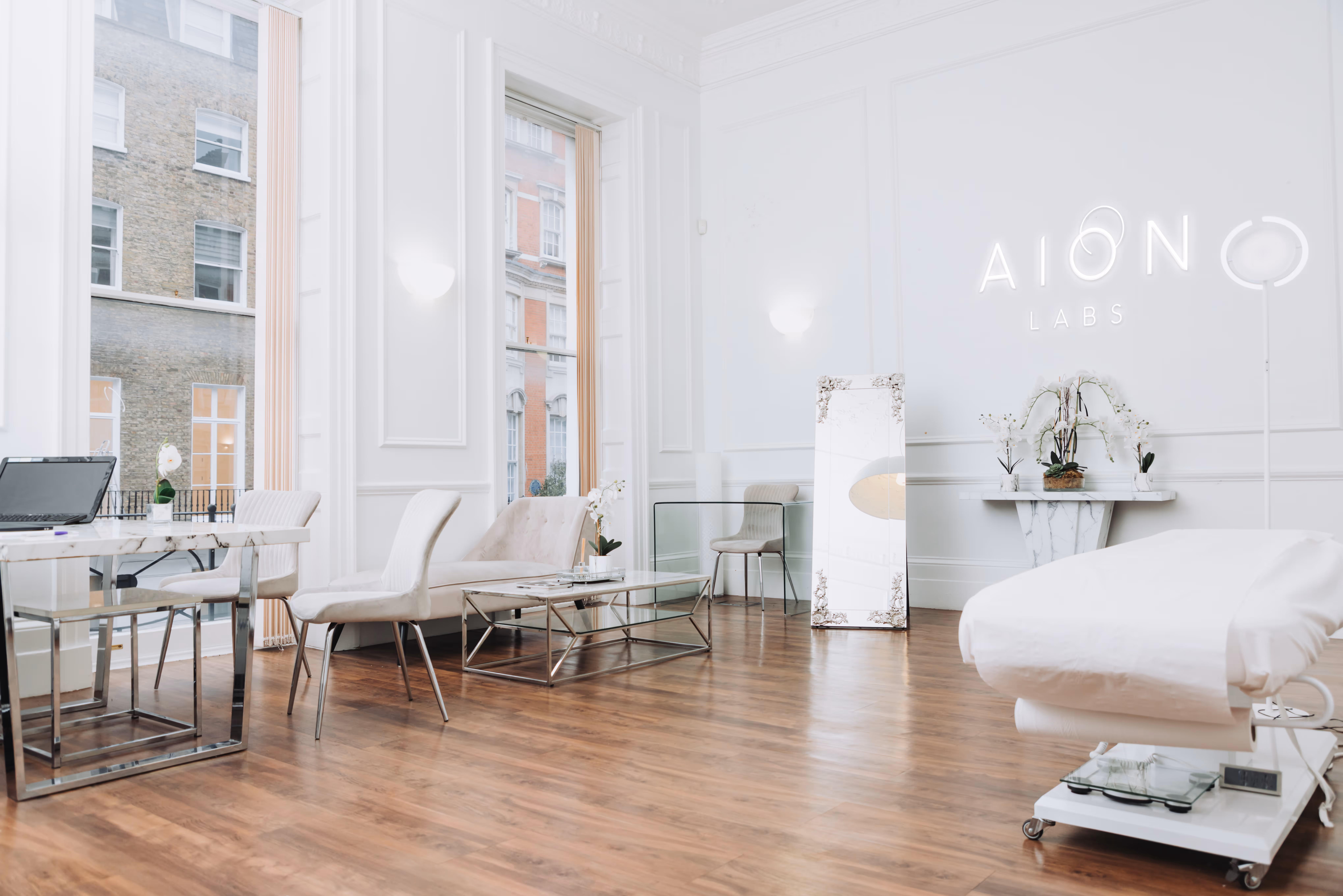 Bright modern clinic room with white walls, wooden floor, lounge chairs, glass table, and a treatment bed under AION Labs neon sign.