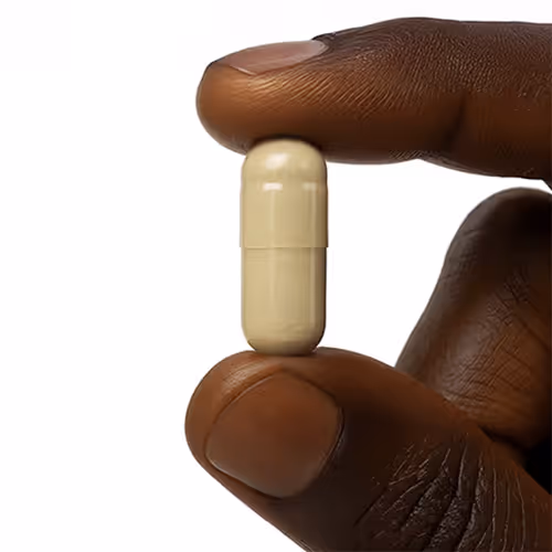 Close-up of a dark-skinned hand holding a beige capsule pill between thumb and forefinger against a white background.