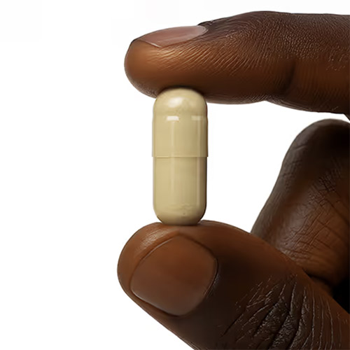 Close-up of a dark-skinned hand holding a beige capsule pill between thumb and forefinger against a white background.