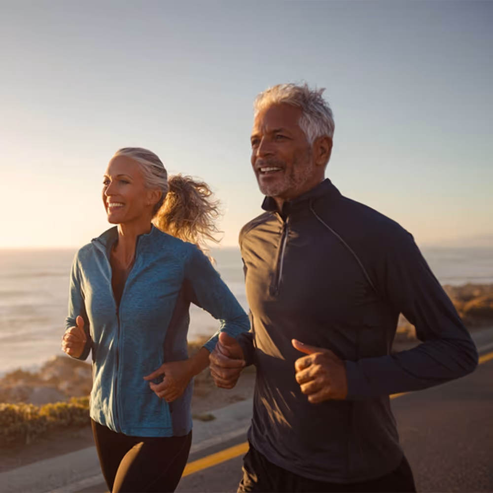 Smiling older man and woman jogging together along a coastal road at sunrise.