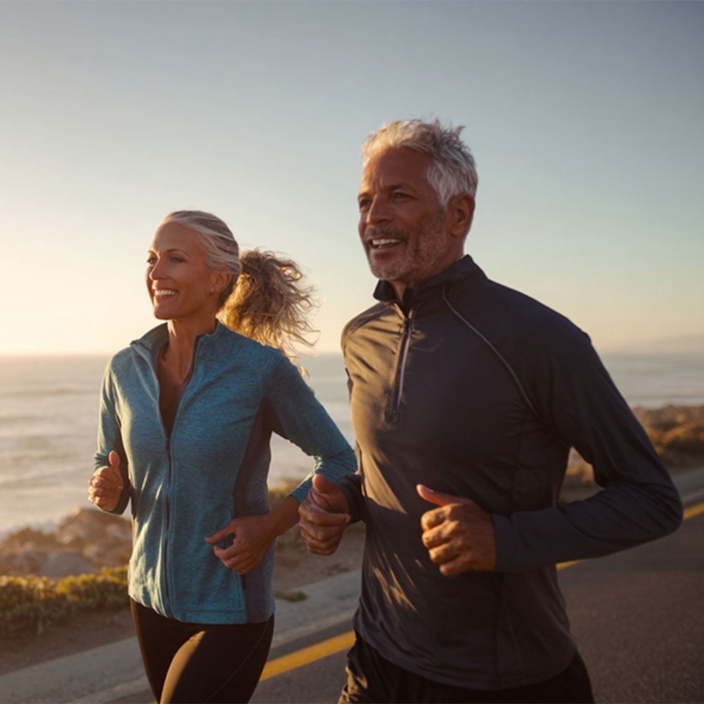 Smiling older man and woman jogging together along a coastal road at sunrise.