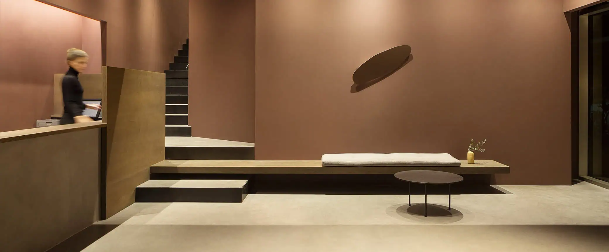 A modern, minimalist interior featuring a wooden bench, round table, and staircase against a warm-toned wall. Soft lighting enhances the ambiance.