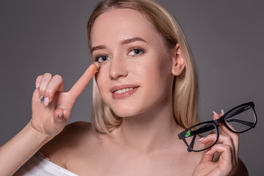 Blonde woman pointing to eye, holding glasses against gray background