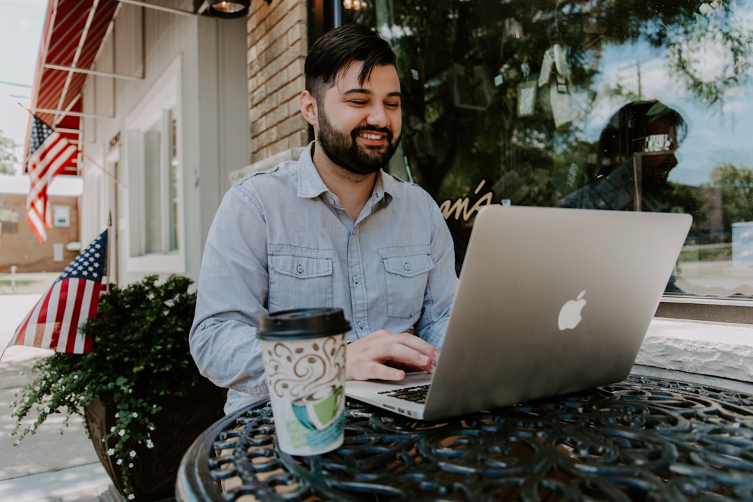 man in gray denim dress shirt smiling while using MacBook Pro in a coffee shop, demonstrating digital marketing strategies for cafes