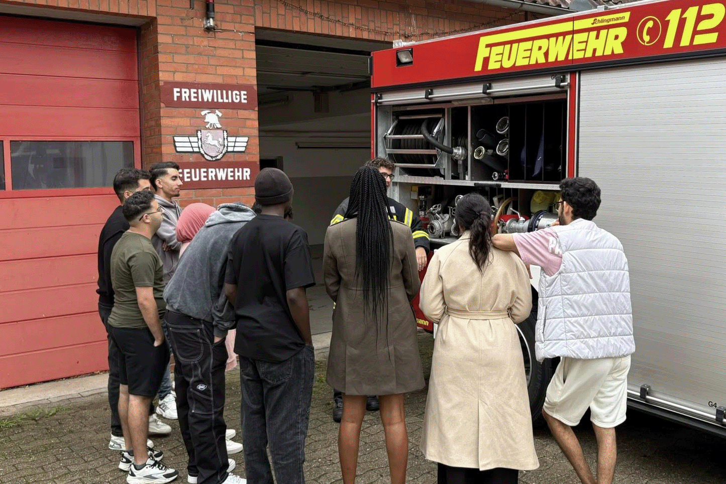 Azubis stehen im Kreis um den Feuerwehrwagen, wärend ein Feuerwehrmann erklärt. Die Seite ist geöffnet und man kann die Ausrüstung sehen.