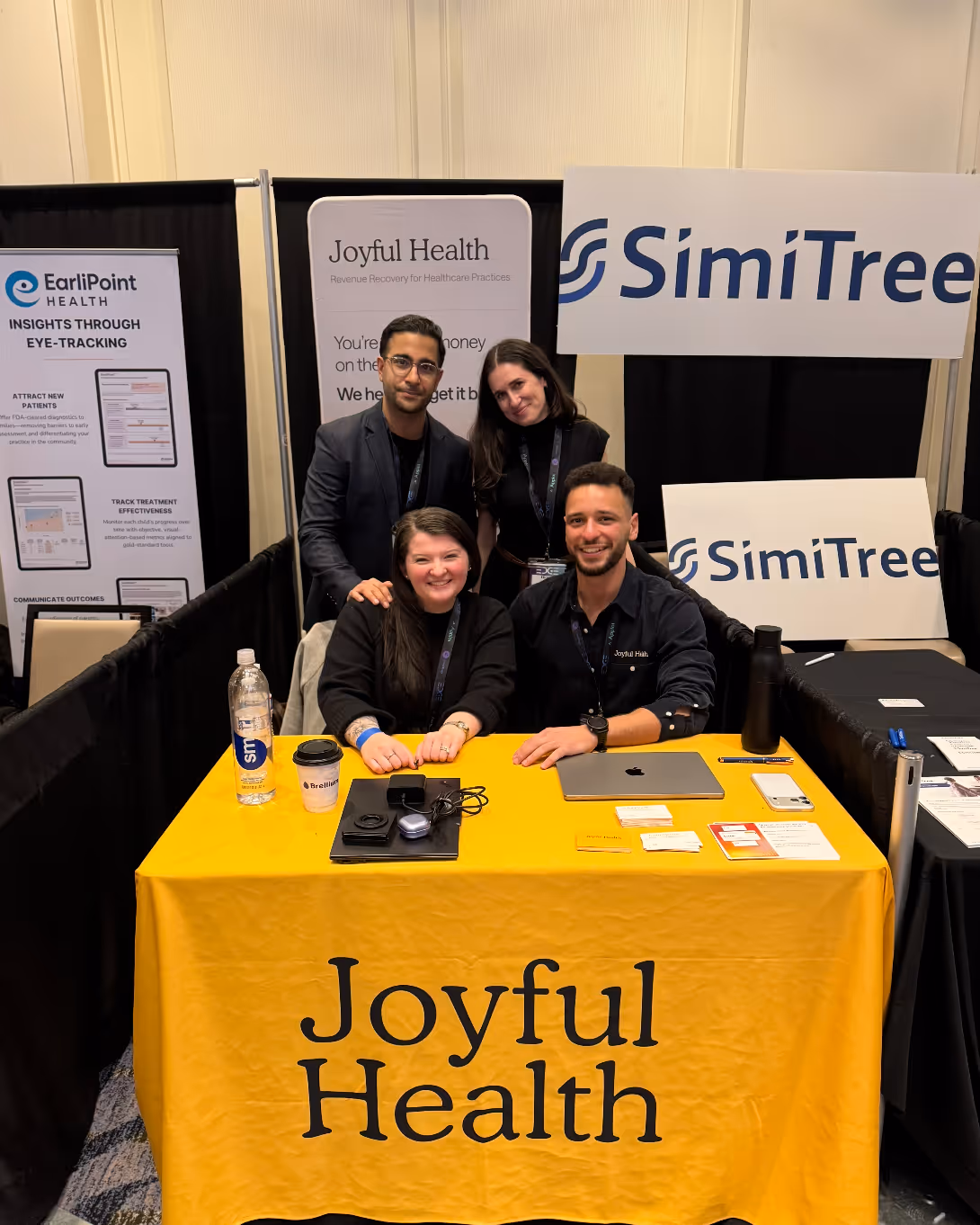 Four people smiling behind a yellow tablecloth with 'Joyful Health' at a booth with banners for Joyful Health, SimiTree, and EarliPoint Health.