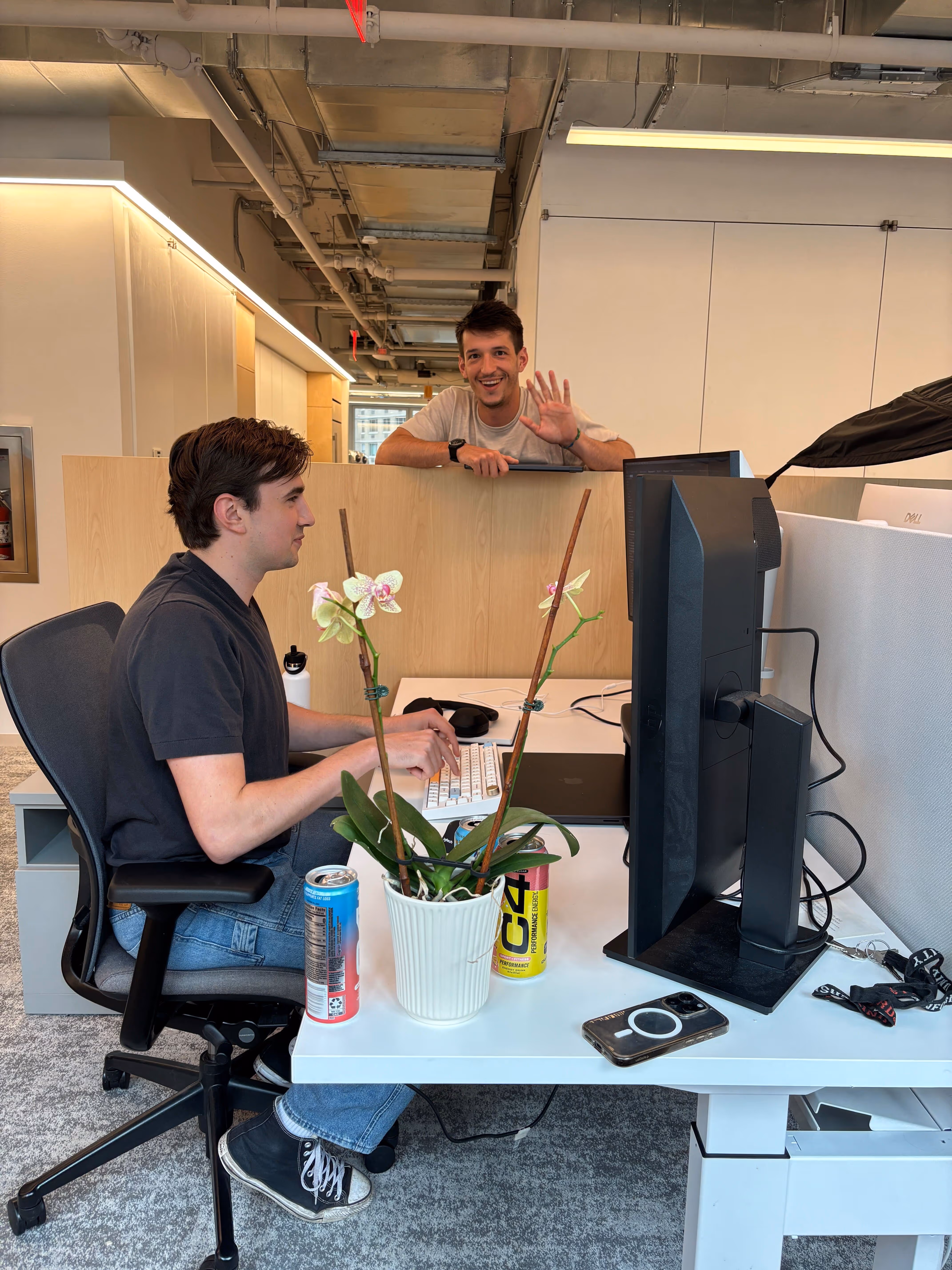 Two men in a modern office space; one sitting at a desk typing on a keyboard and the other leaning on a divider waving and smiling.