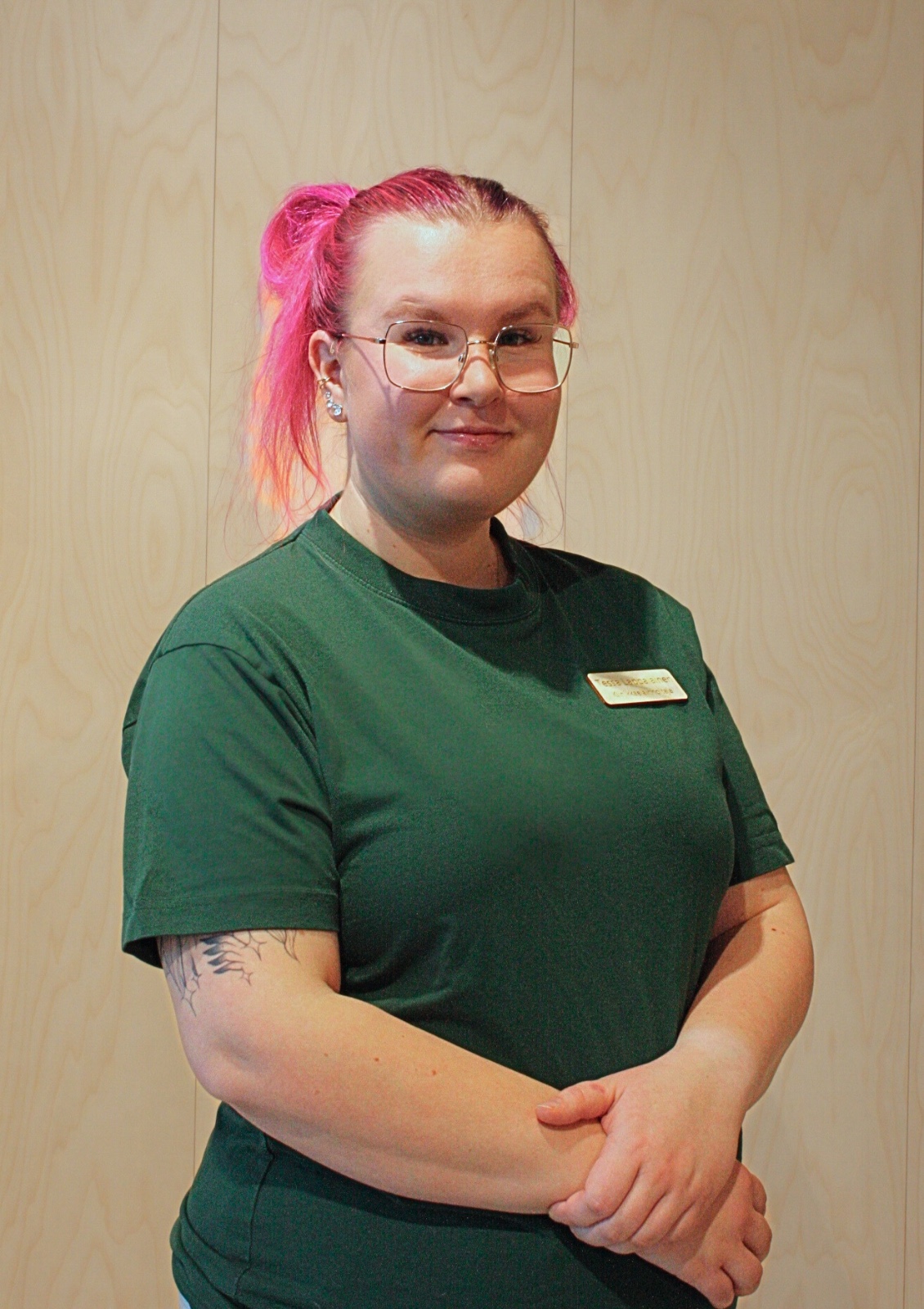 Kuva naisesta, jolla on vaaleanpunaiset hiukset, silmälasit ja vihreä paita, seisomassa puupaneeliseinää vasten. Hänellä on kädet ristissä ja hän hymyilee. Valo on pehmeää ja tasaista.