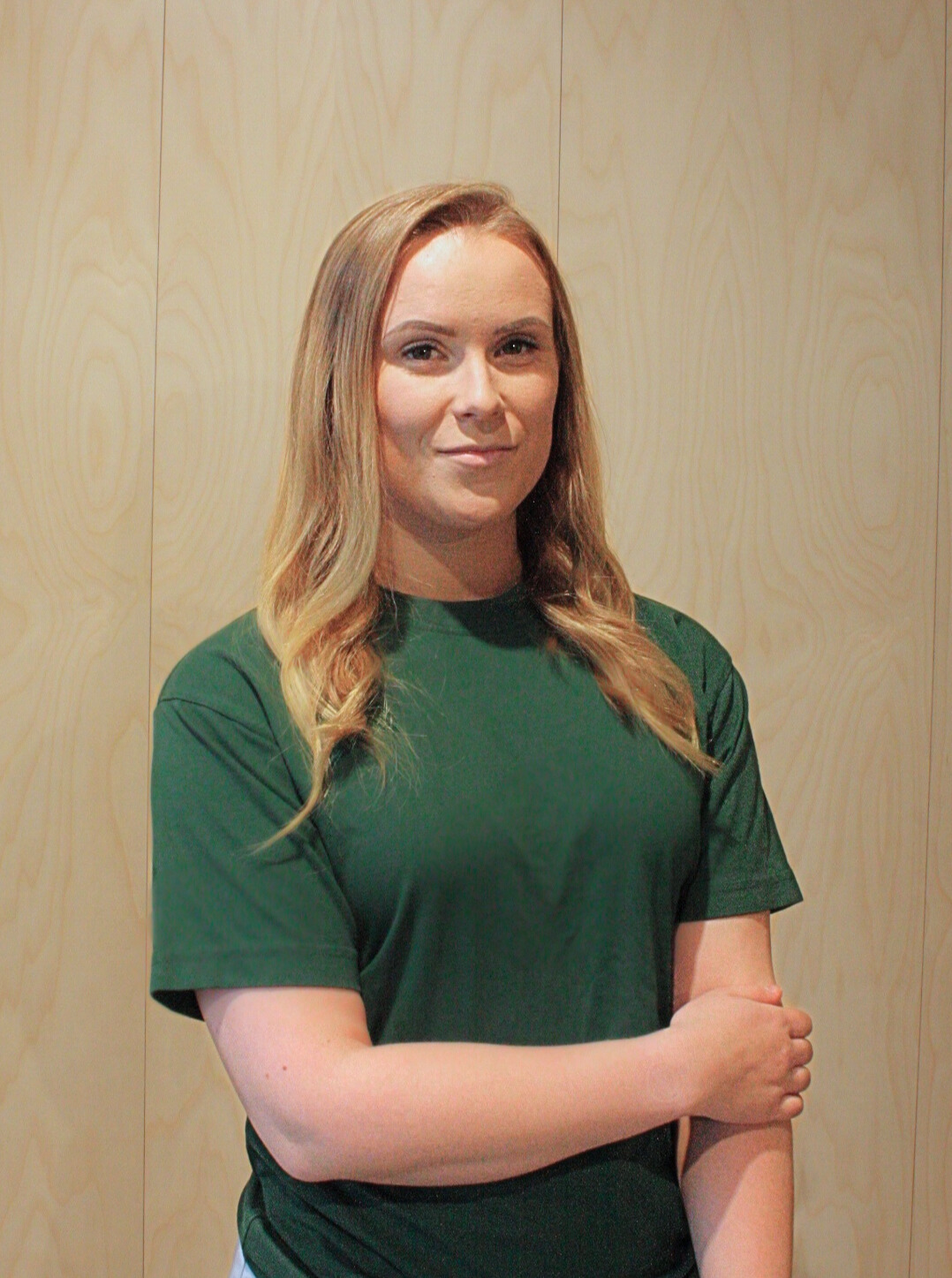 Vaaleahiuksinen nainen vihreässä paidassa seisoo puupaneeliseinää vasten, kädet ristissä.