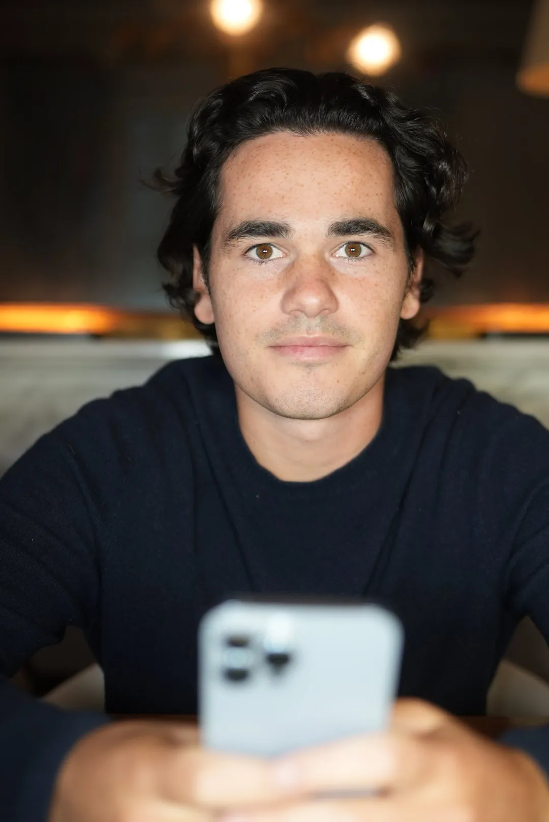 Young man with dark hair and brown eyes wearing a dark sweater, holding a smartphone and looking at the camera.