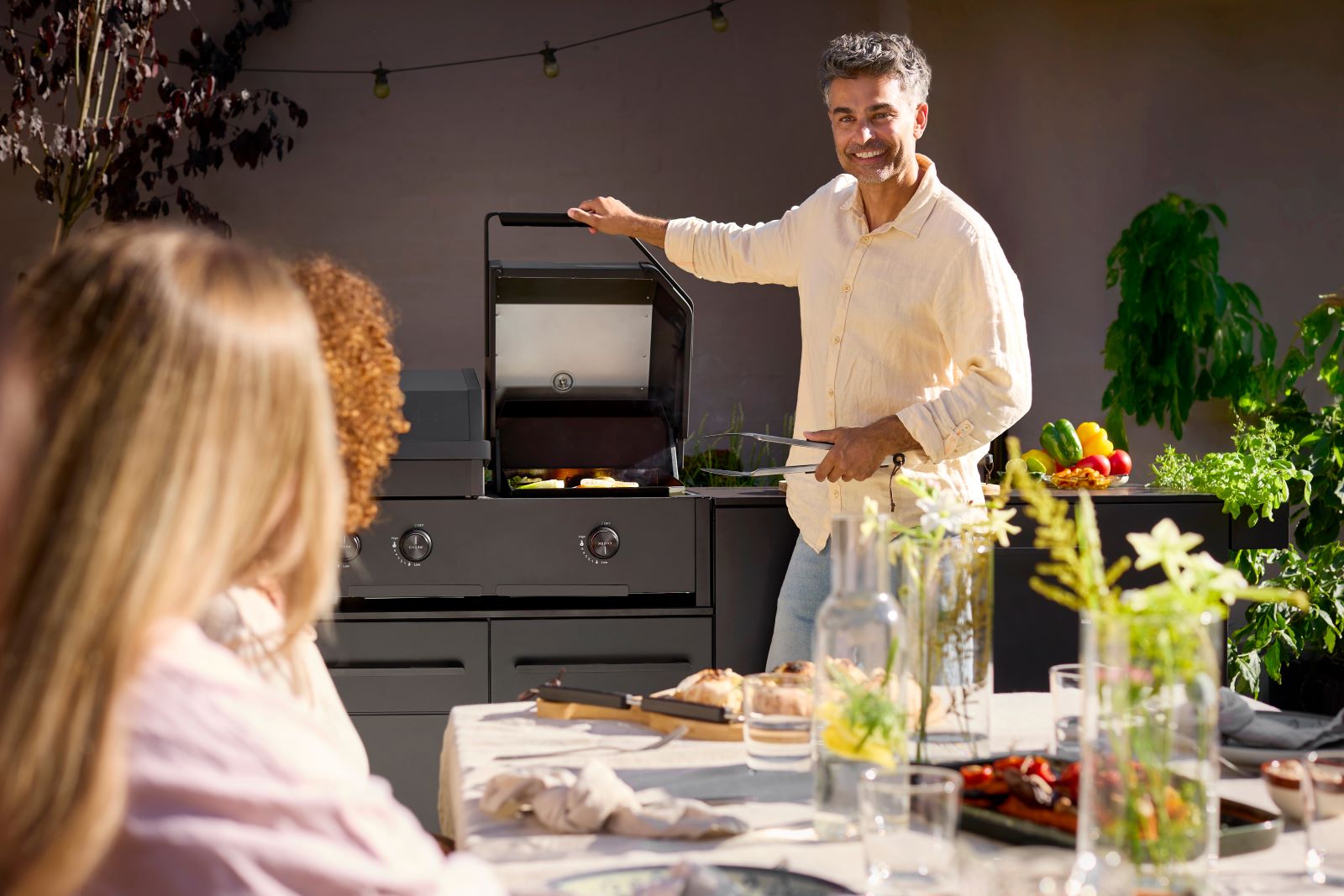 Mann in einem hellen Hemd grillt Gemüse auf einem Grill, während zwei Personen an einem mit Speisen und Getränken gedeckten Tisch im Freien zusehen.