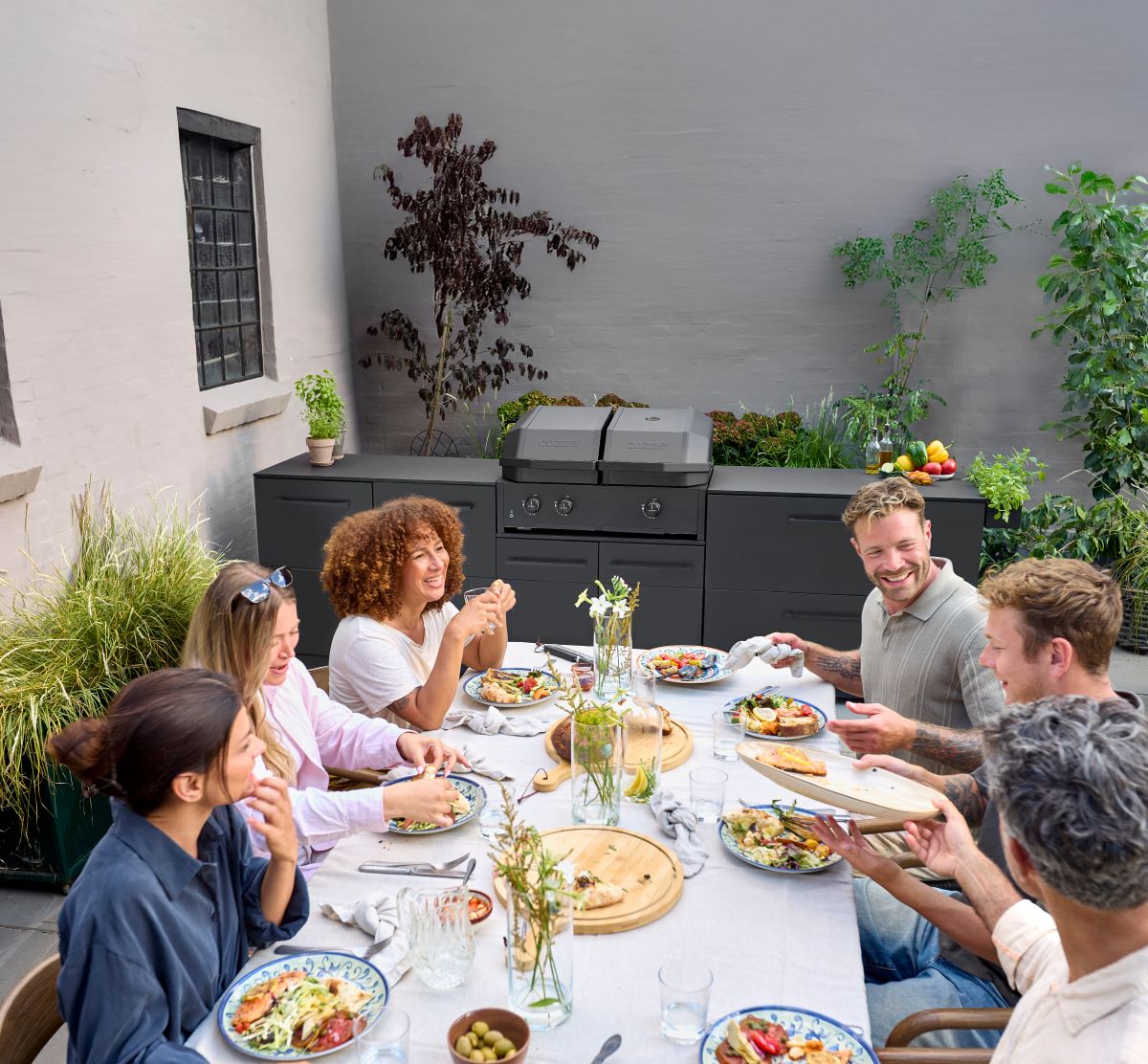 Eine Gruppe von sechs Freunden genießt ein Essen im Freien an einem Tisch mit Tellern voller Speisen und einem modernen Grill im Hintergrund.