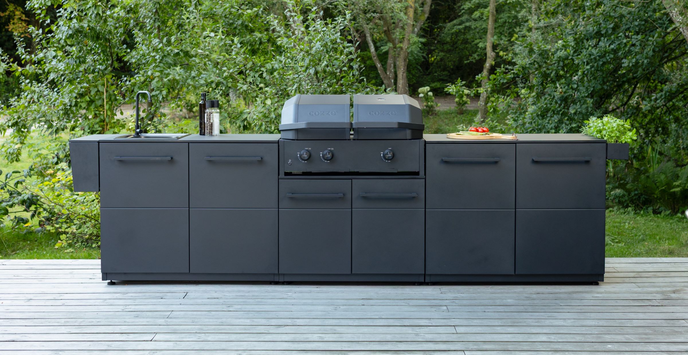 Moderne schwarze Außenküche mit eingebautem Grill, Spüle und Kräutern auf einer Holzterrasse, umgeben von viel Grün.