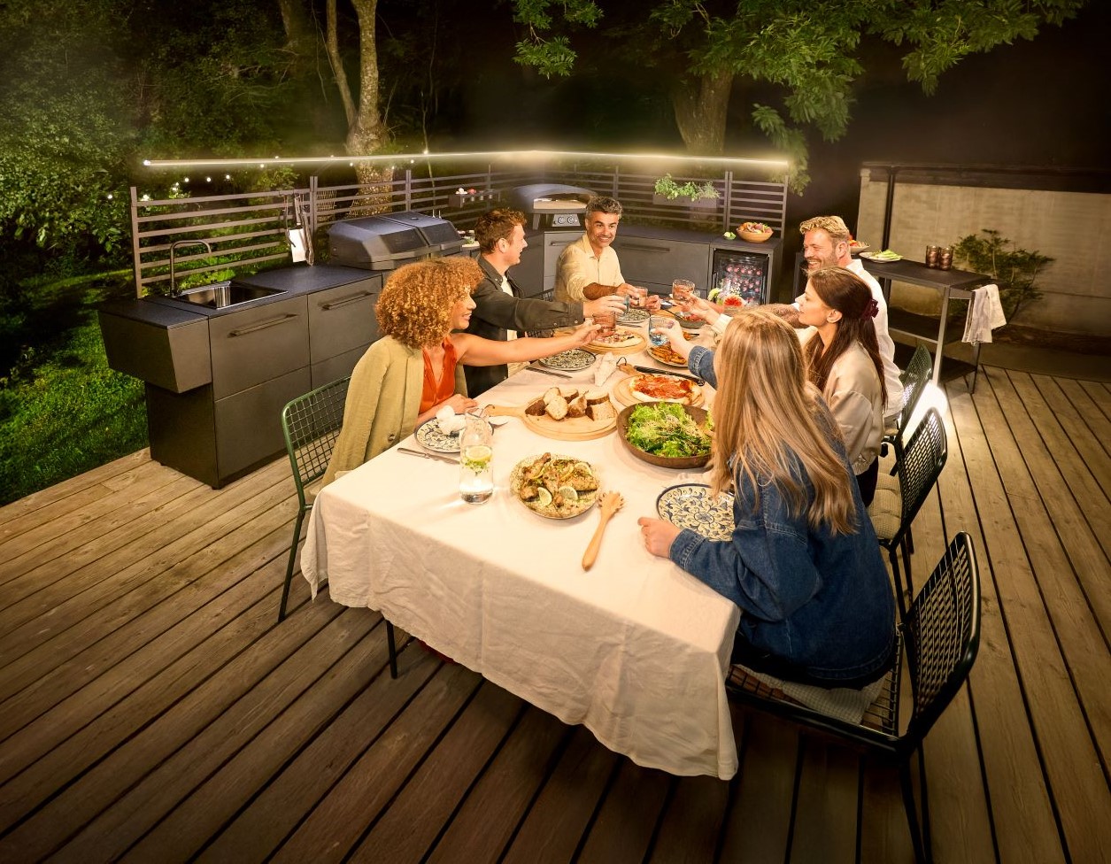 Sechs Freunde stoßen abends in einer warm beleuchteten Außenküche mit Essen an einem Tisch mit Getränken an.