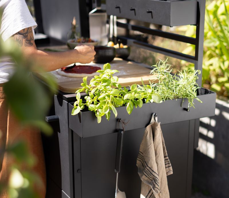 Person, die auf einer schwarzen Kochstation im Freien Essen zubereitet, vor der frische grüne Kräuter gepflanzt sind und an einem Haken ein Handtuch hängt.