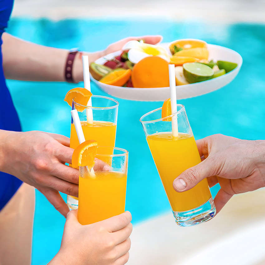 Hands holding glasses of orange juice with fruit platter by the pool, celebrating summer with refreshing drinks