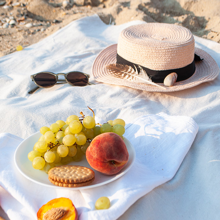 Beach picnic setup with fruit, cookies, sunglasses, and a straw hat on a blanket—perfect for Orderly’s beach-ready snack delivery service