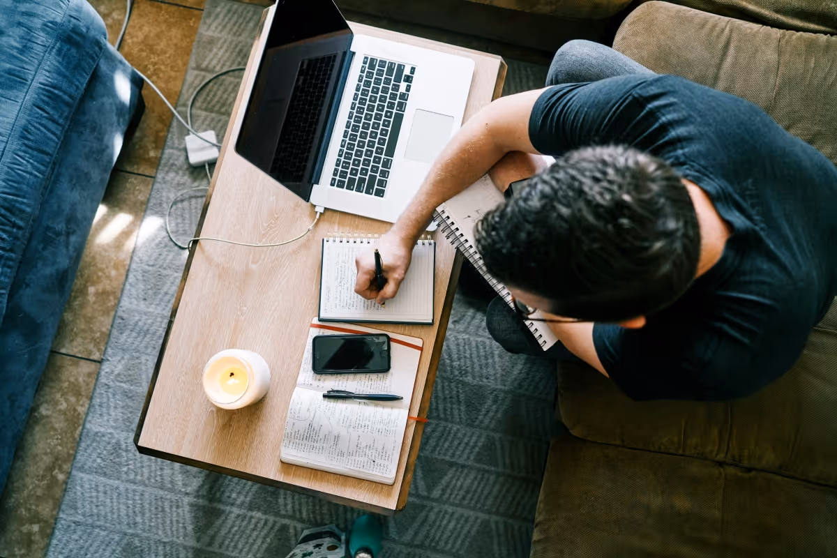 Eine Person sitzt von oben gesehen am Couchtisch, macht sich Notizen in ein Notizbuch, vor ihr ein Laptop und eine brennende Kerze.