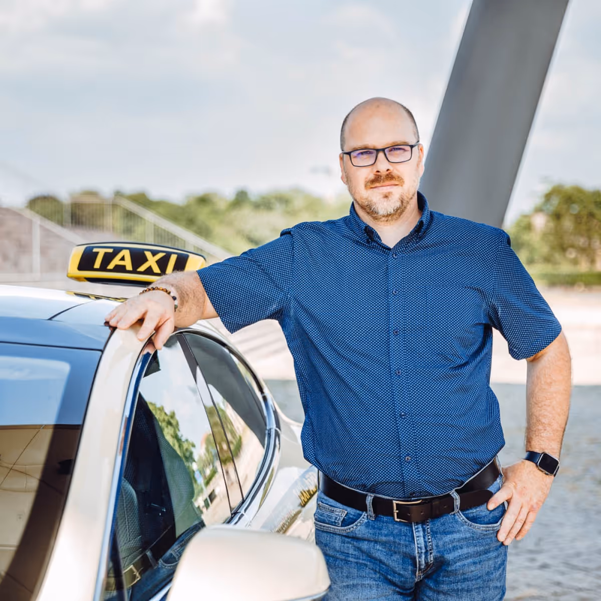 Ein lächelnder Taxifahrer mit Brille und Bart lehnt am offenen Fenster seines Taxis.
