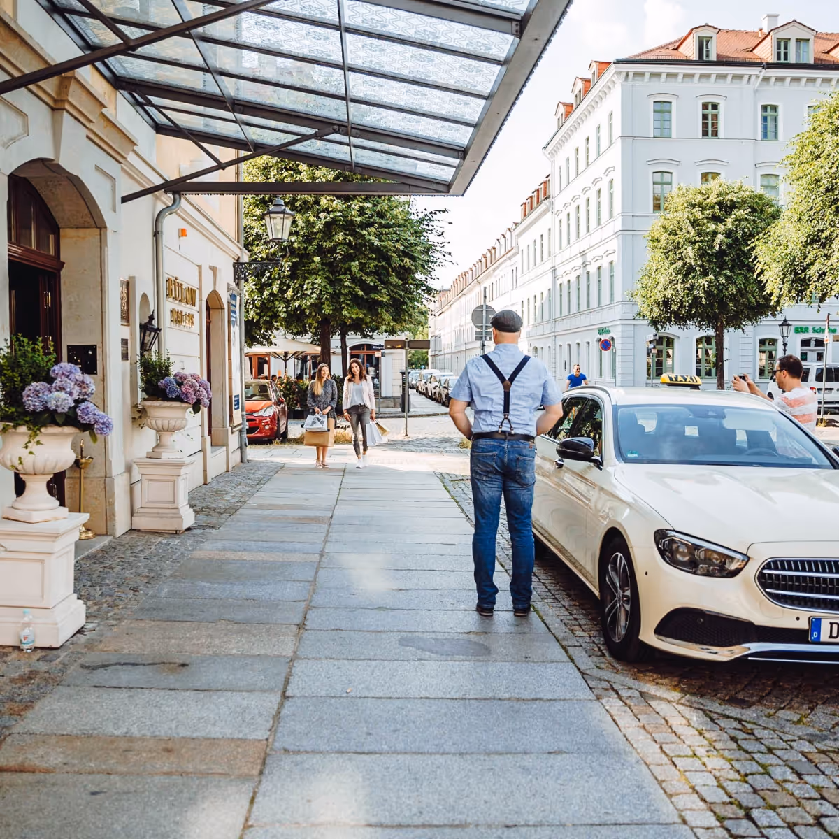 Ein Taxifahrer steht an seinem geparkten Auto und wartet auf Fahrgäste, die aus einem Hotel kommen.
