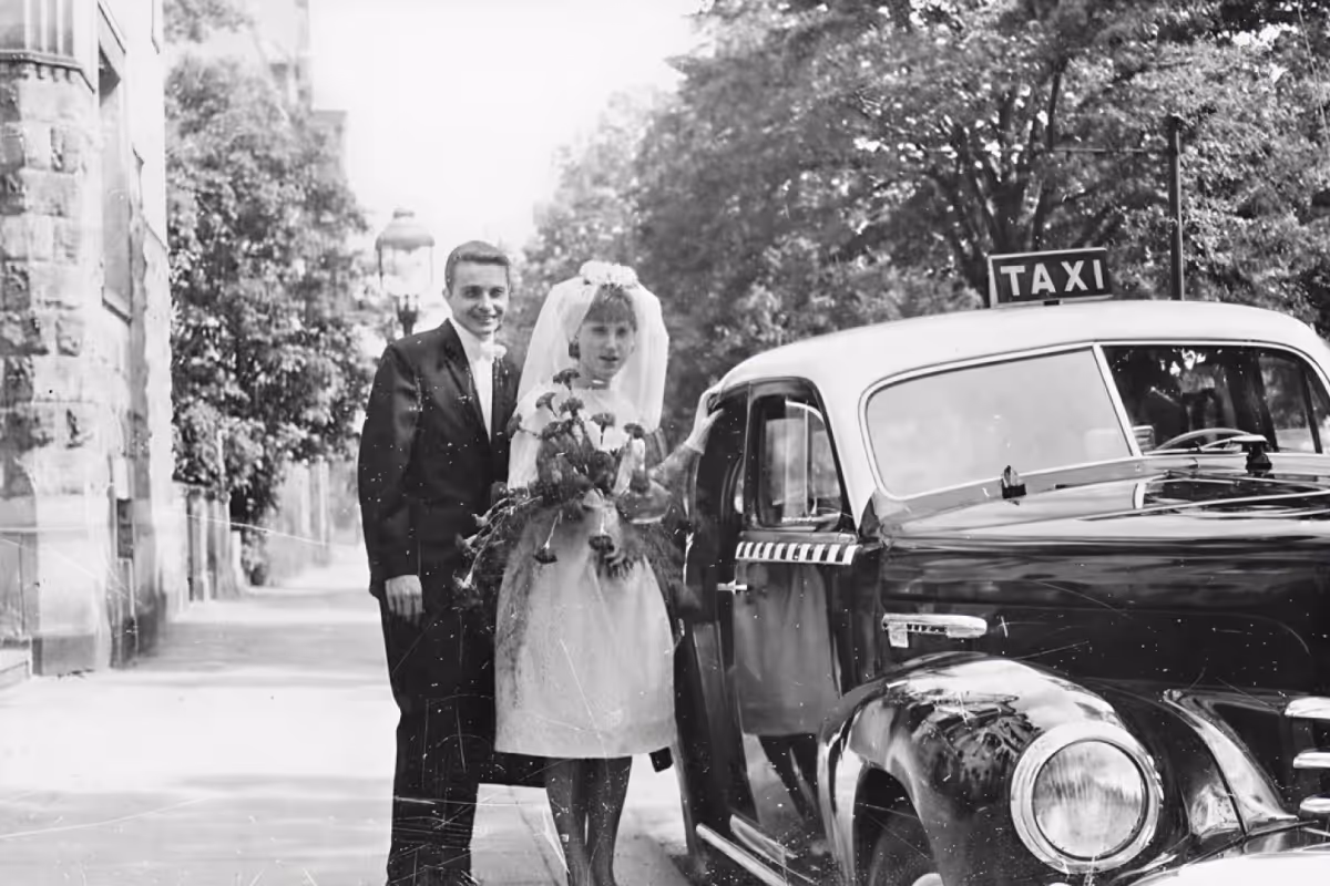 Ein Schwarz-Weiß-Foto eines Brautpaares, das neben einem historischen Taxi steht.

