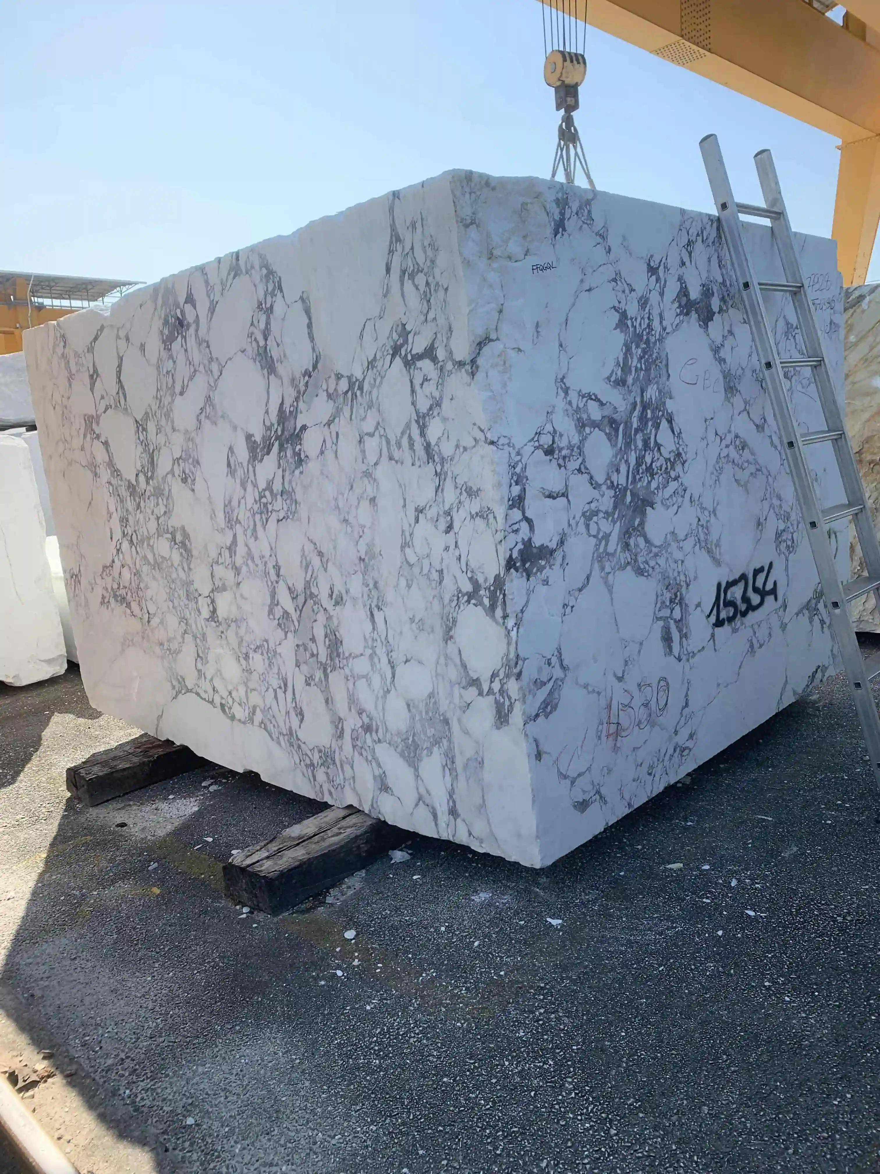 Large block of white marble with grey veining supported on wooden beams, with a metal ladder leaning against it and a crane hook above.