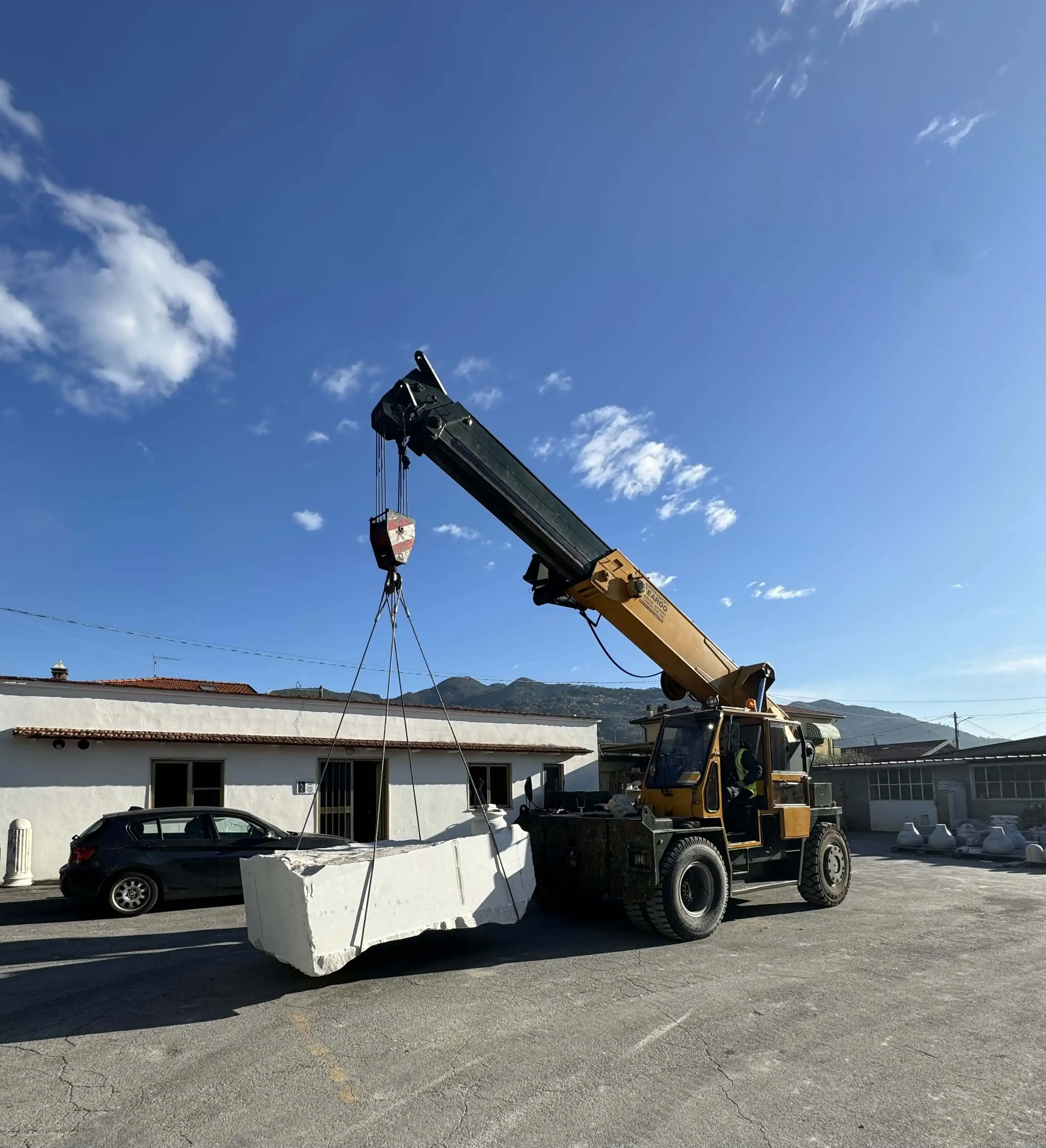 Yellow crane lifting a large rectangular white stone block in an outdoor yard with buildings and mountains in the background.