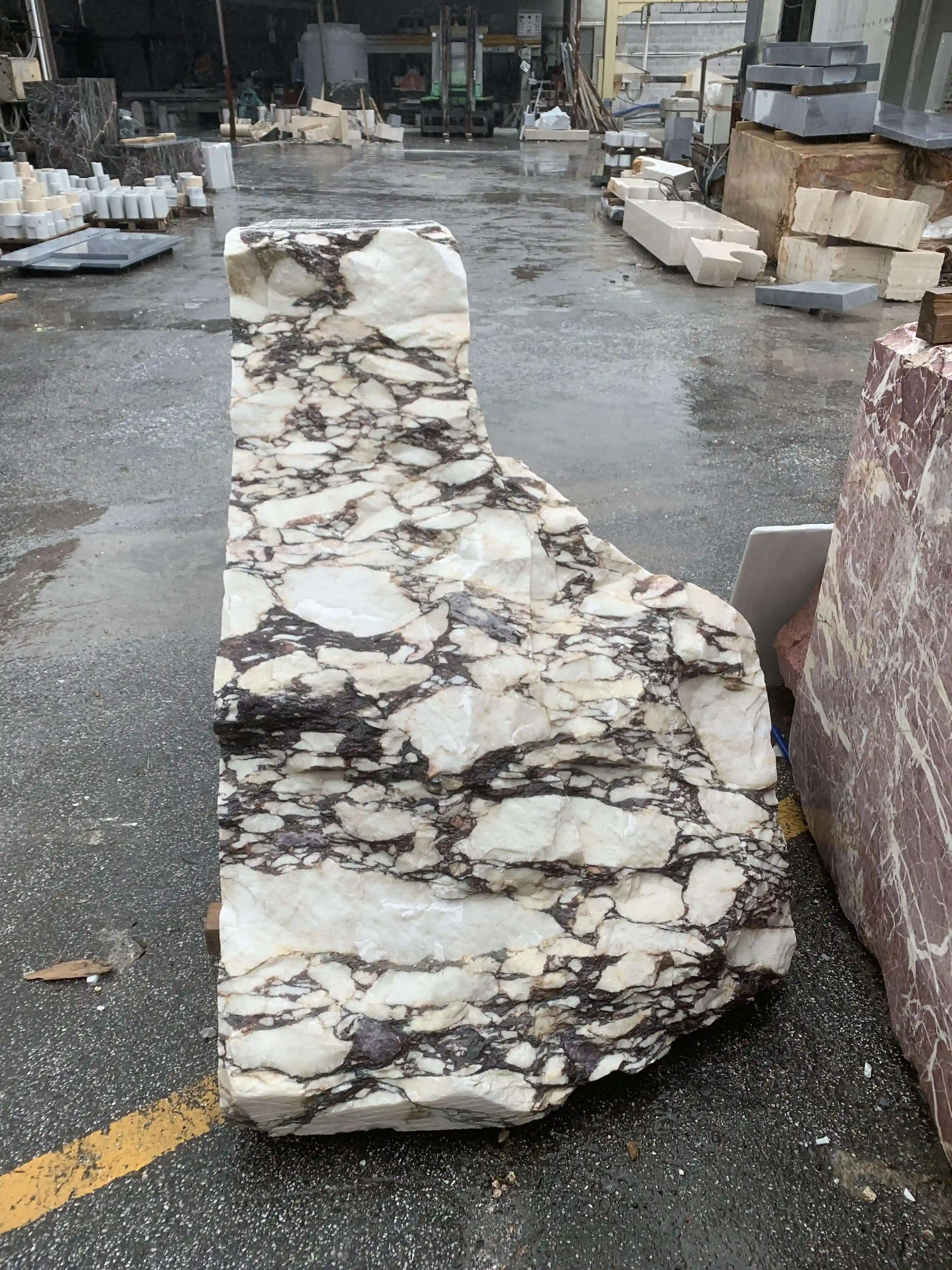 Large irregularly shaped block of white marble with dark brown veining sitting on wet pavement in a stone yard.