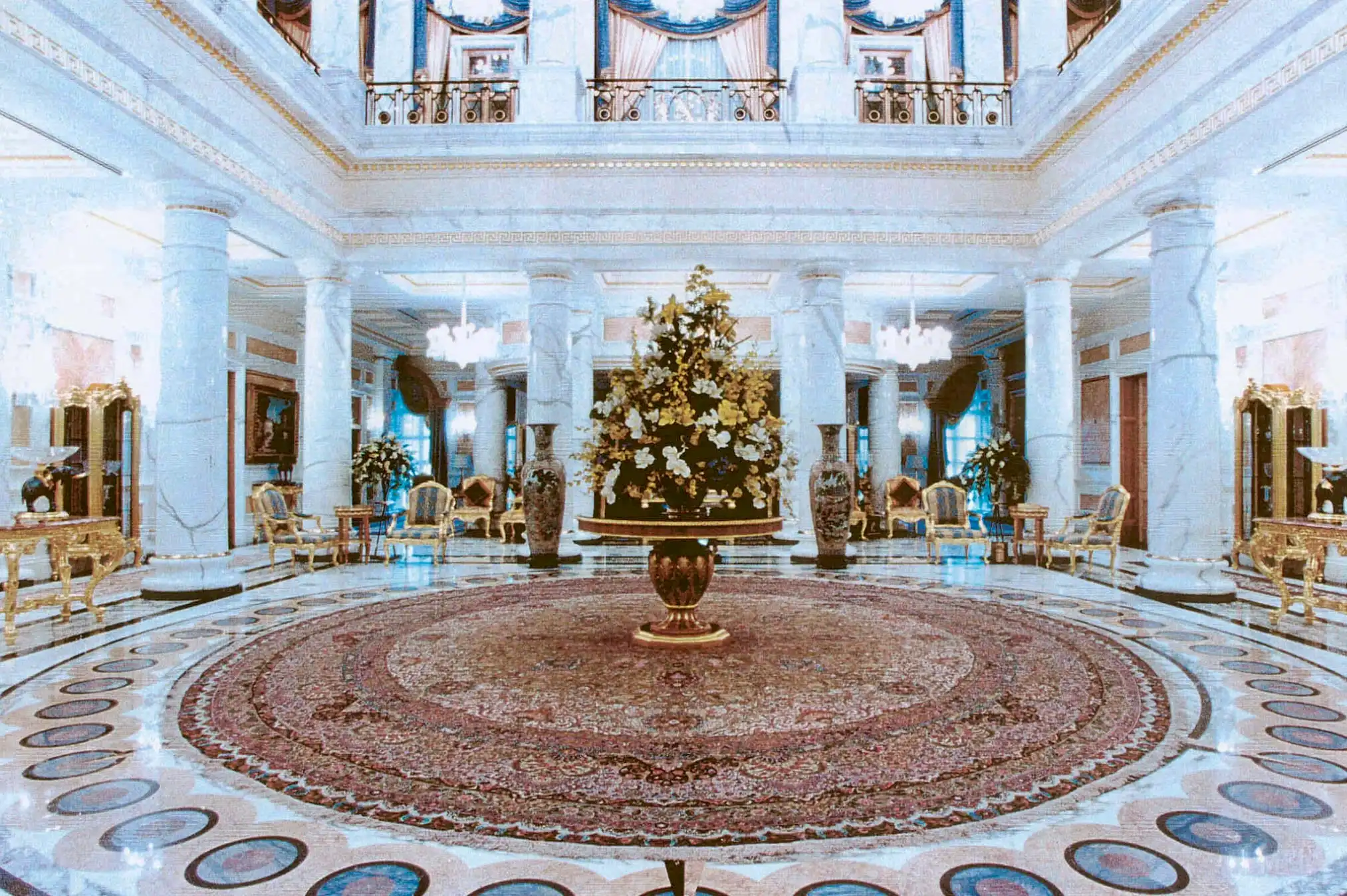 Luxurious marble foyer with large ornate round rug, central floral arrangement on a pedestal, white columns, and elegant gold-trimmed furniture.