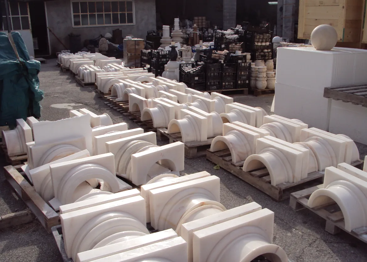 Stacks of white marble architectural elements arranged on wooden pallets outdoors in a workshop or storage area.
