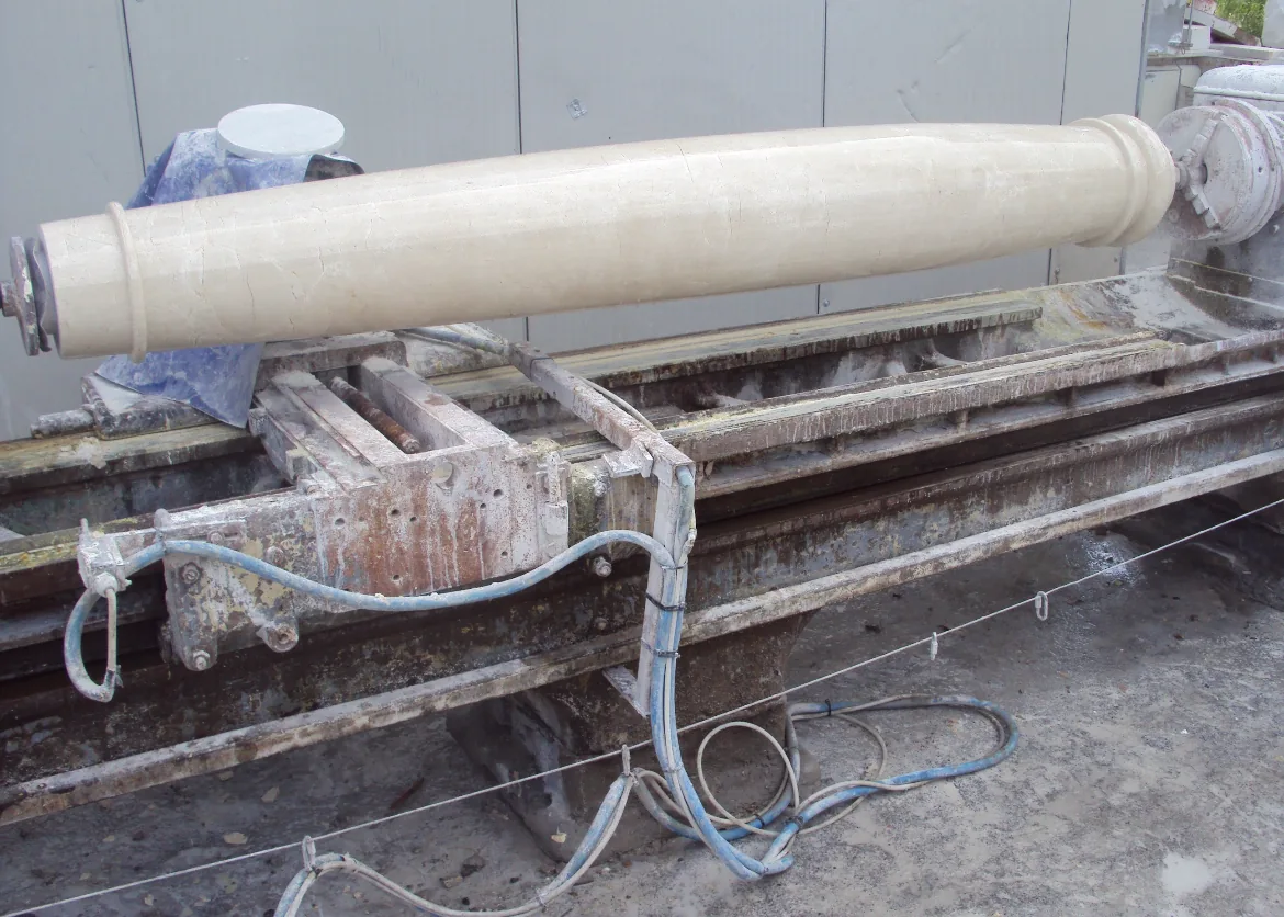 Large cylindrical stone column being shaped on an industrial lathe machine surrounded by hoses and dust.