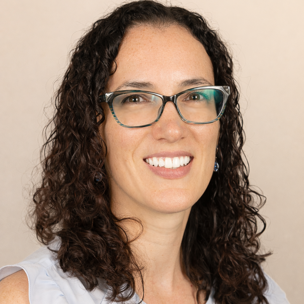 Smiling woman with curly dark hair and glasses wearing a light blue sleeveless blouse against a neutral background.