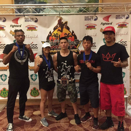 Five people pose proudly in front of a martial arts championship banner. Three wear medals, with a mix of casual and athletic clothing, conveying triumph.