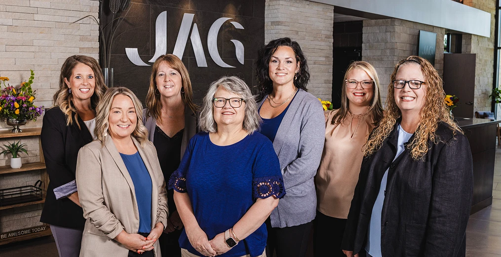 Seven team members from Jones Advisory Group standing inside their office in front of their logo sign, smiling.