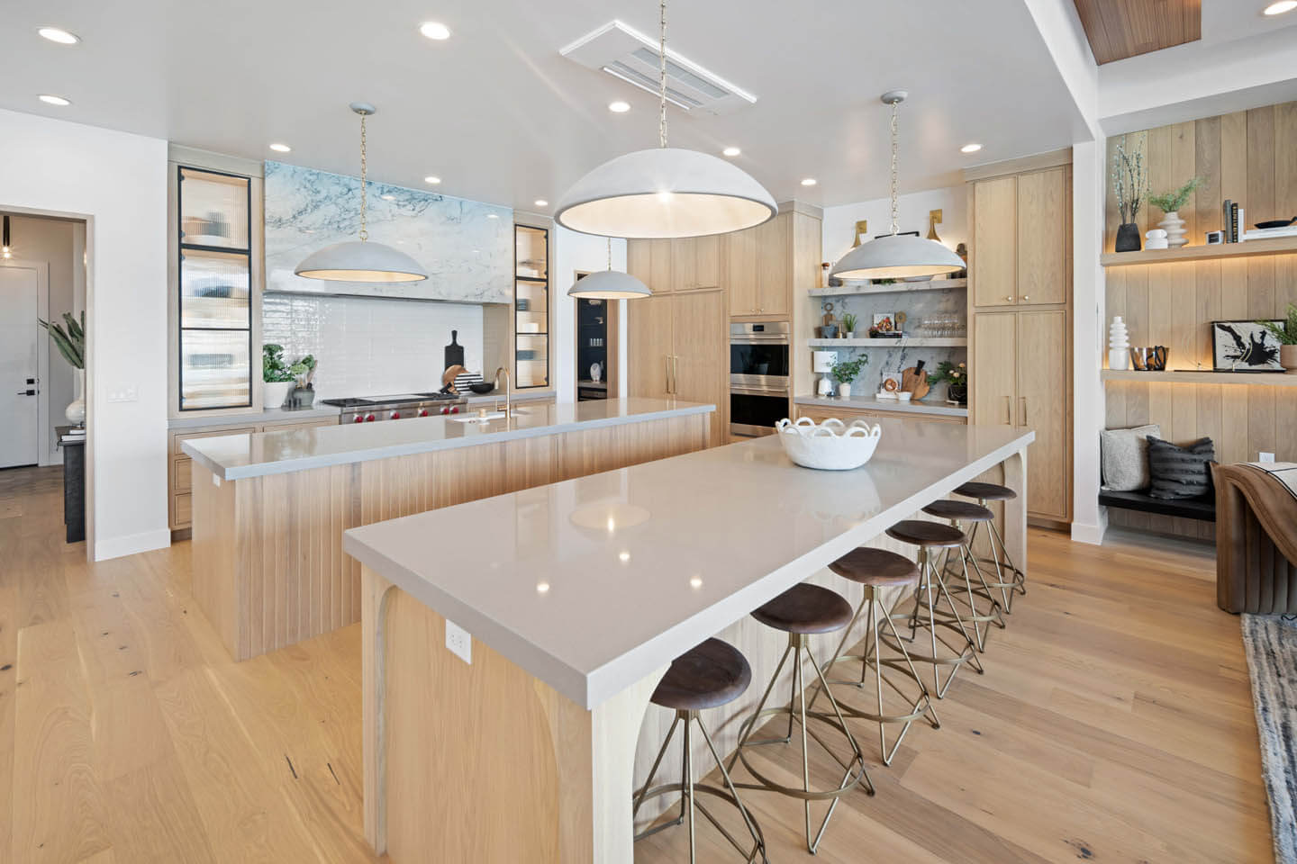 Modern kitchen with two large light-colored islands, six dark cushioned stools, pendant lights, wood cabinets, and open shelving with decor.