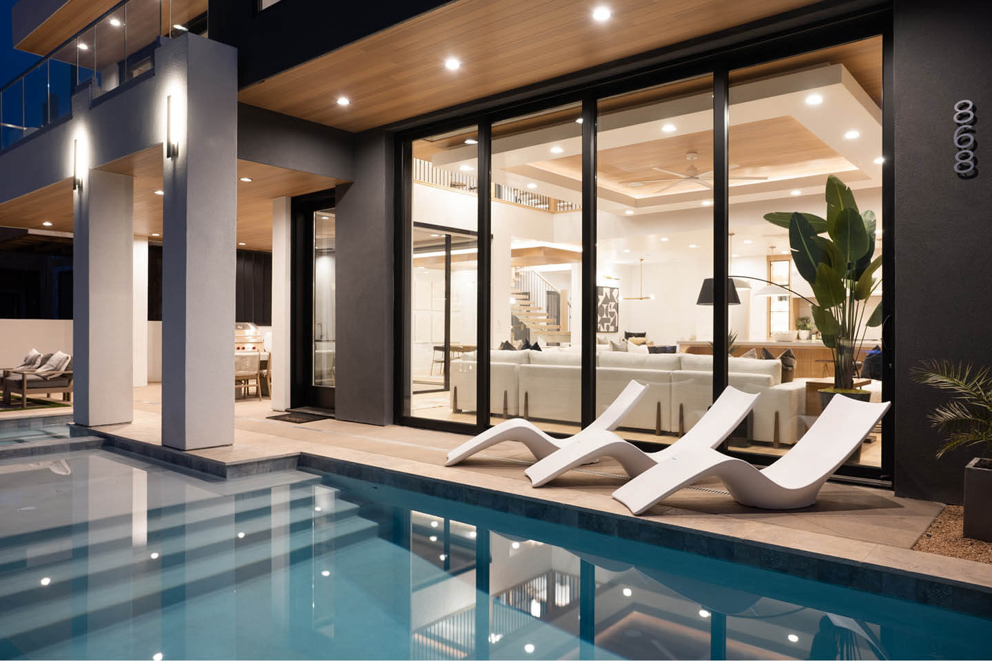 Modern house exterior at night with illuminated large glass windows, a swimming pool, and three white lounge chairs on the poolside patio.