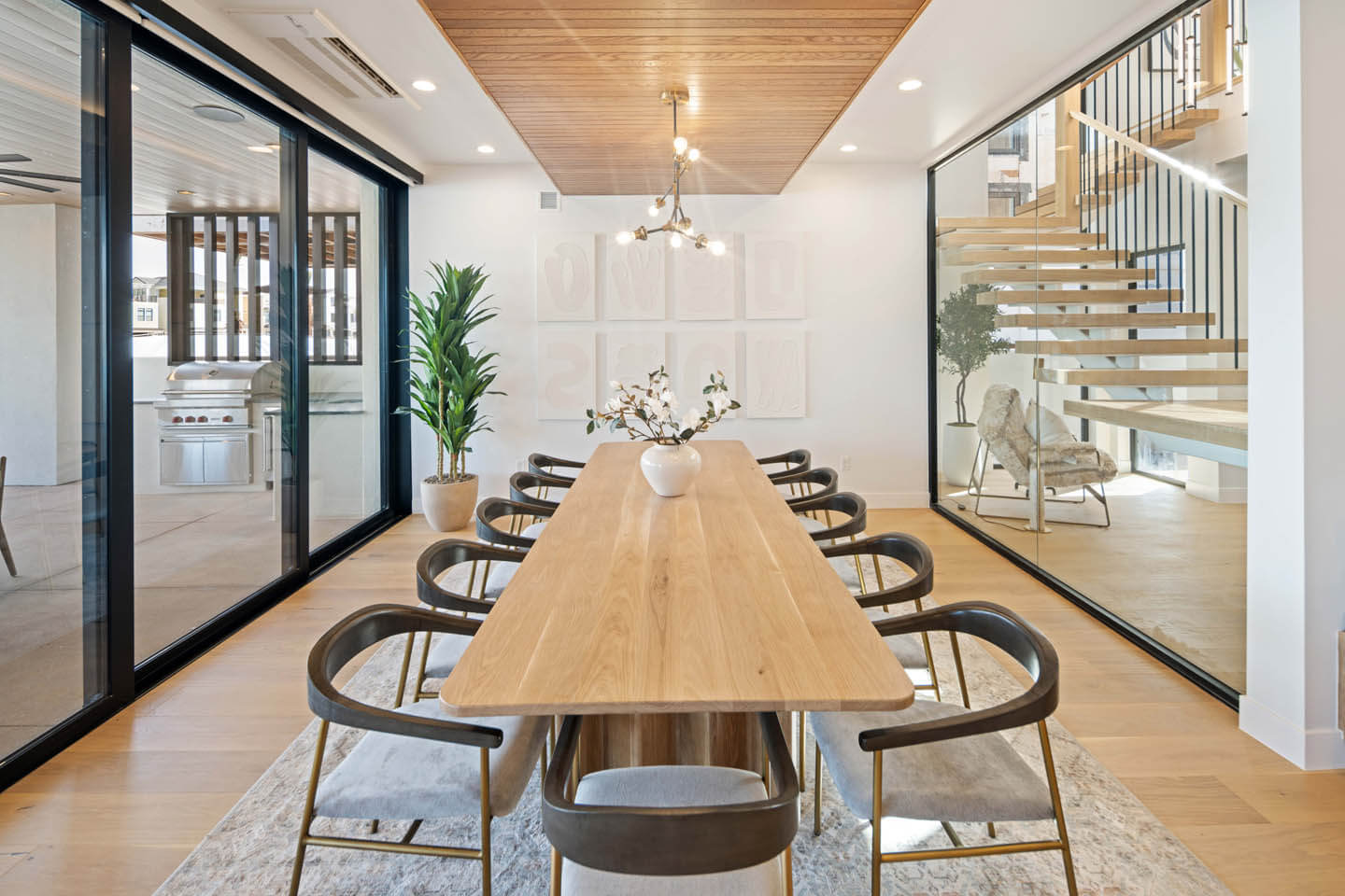 Modern dining room with a long wooden table, eight chairs, a white vase with flowers, large glass doors, and a floating wooden staircase.